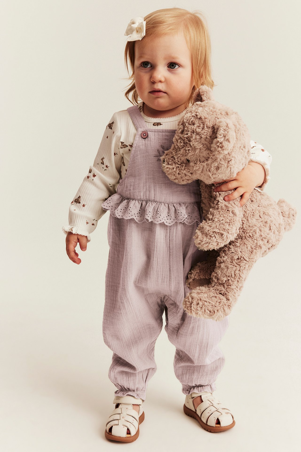 Framifrån: Baby bär ljuslila bomullshängselbyxa med spets, över mönstrad tröja, hållande en brun nalle.