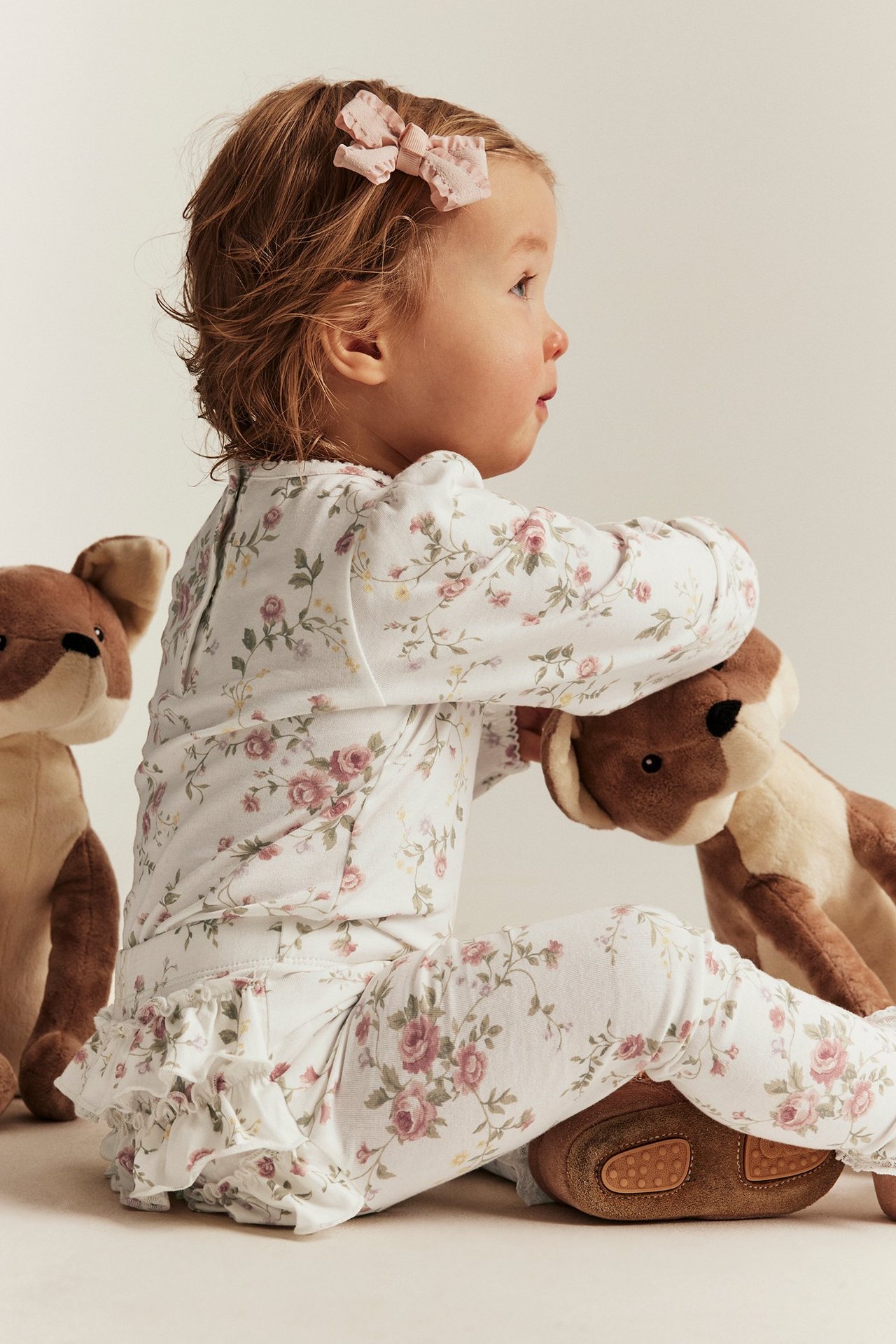 Sidebilde av baby i offwhite blomstrete rysje-leggings, matchende topp, rosa sløyfe og brune sko.