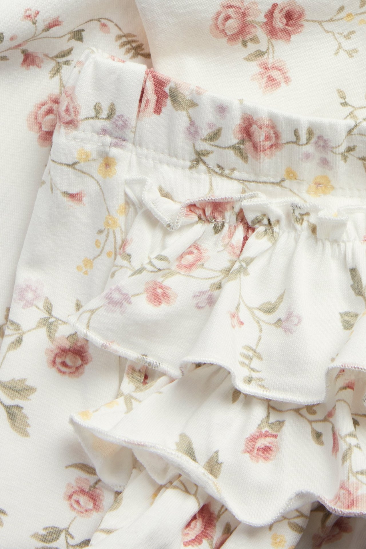 Nærbilde av offwhite baby-leggings med rosa blomstermønster, elastisk midje og tre volanger bak.