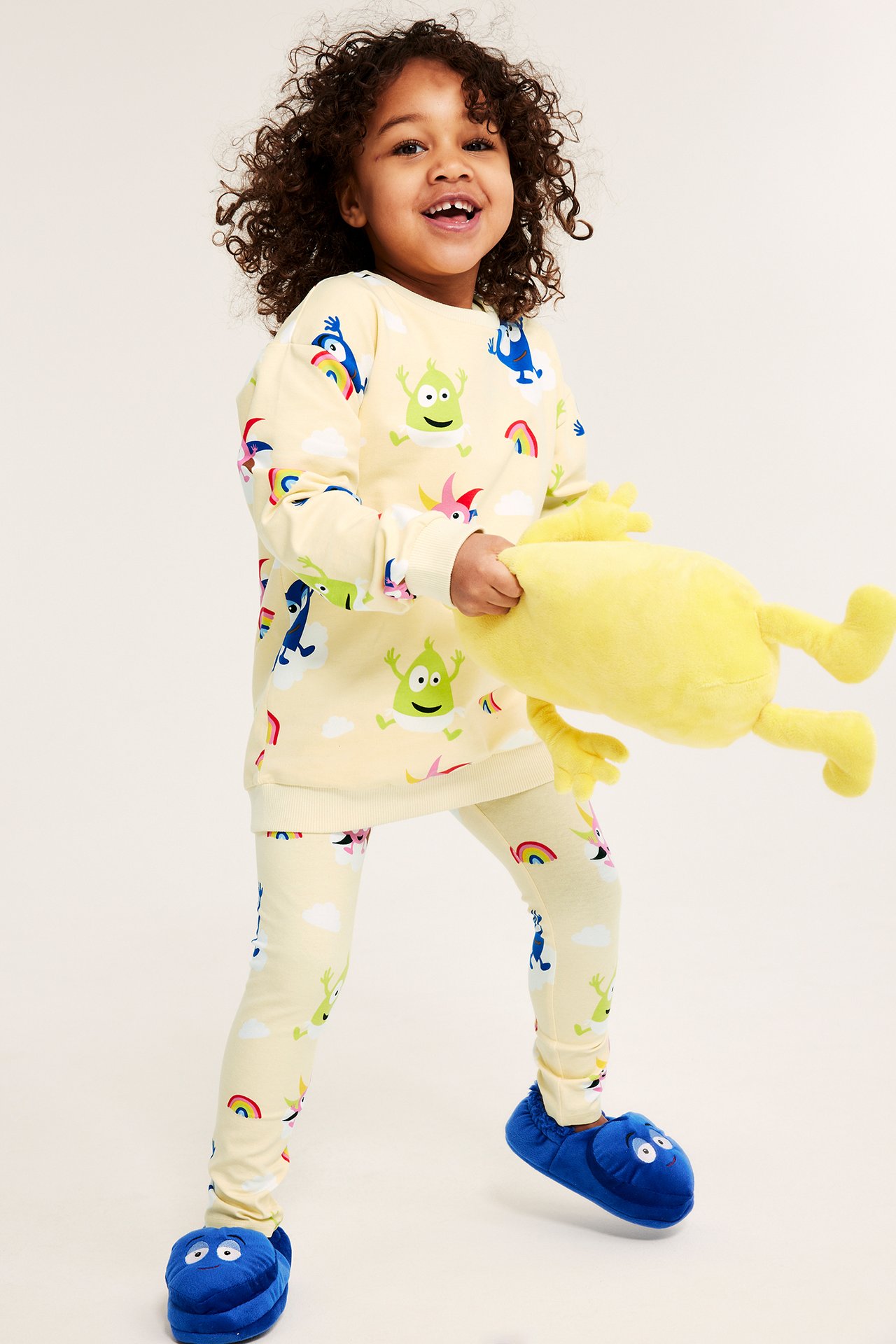 Hymyilevä lapsi Babblarna-leggingseissä ja -paidassa, sinisissä tossuissa, pitää keltaista pehmolelua.