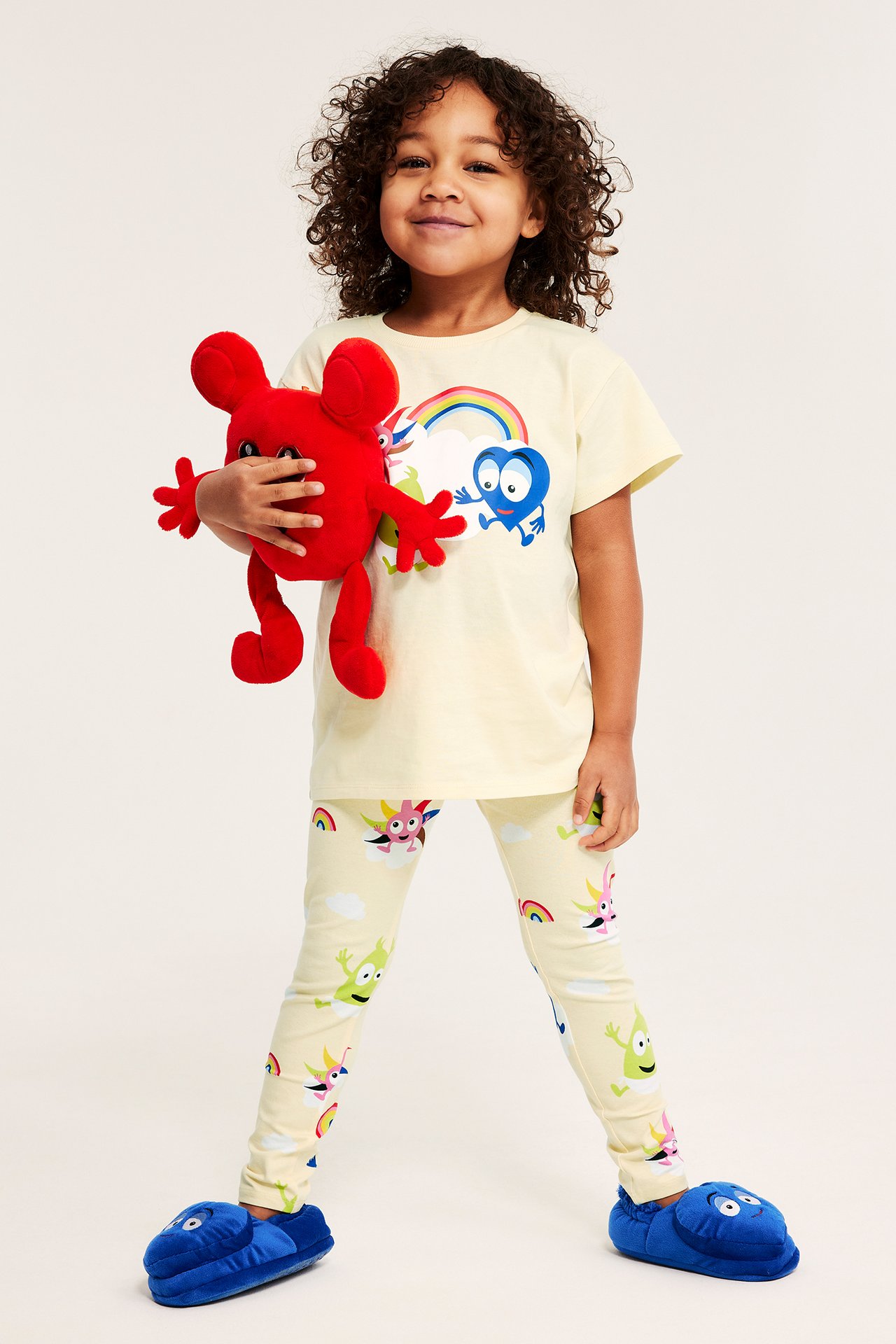 Edestä: Lapsi vaaleanbeigeissä Babblarna-kuvioleggingseissä, keltaisessa t-paidassa, sinisissä tohveleissa, pitelee punaista lelua.