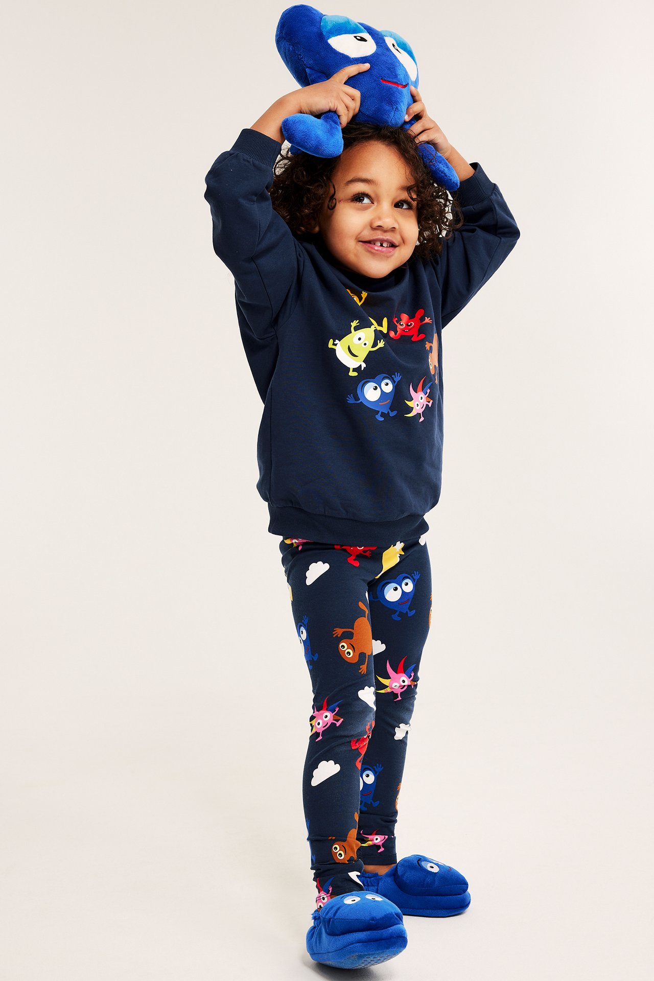 Front: Barn bär mörkblå Babblarna-mönstrade leggings, matchande tröja, blå tofflor, håller mjukdjur.