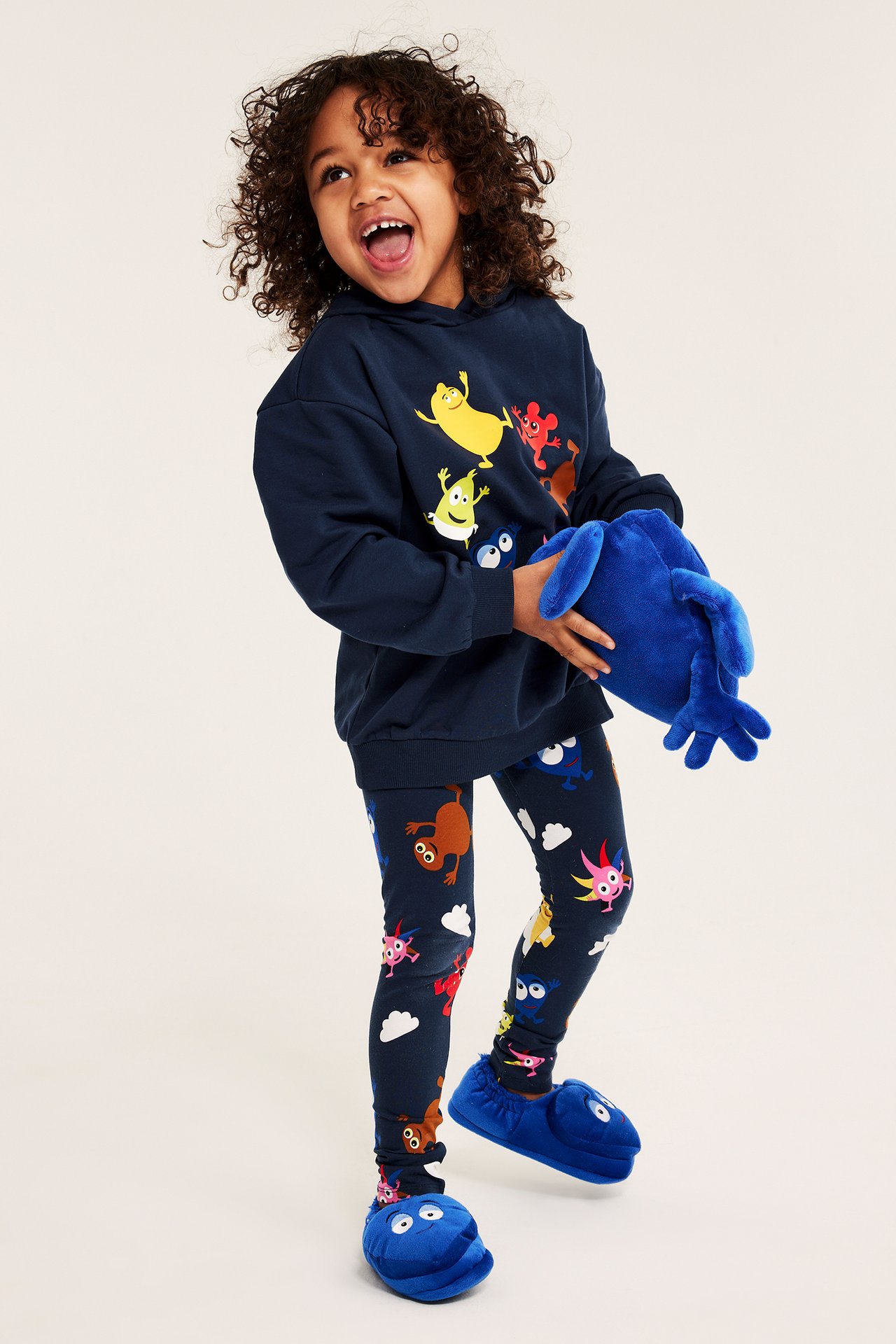 Framifrån: Barn i mörkblå Babblarna-mönstrade leggings, matchande huvtröja, blå tofflor, håller mjukdjur.