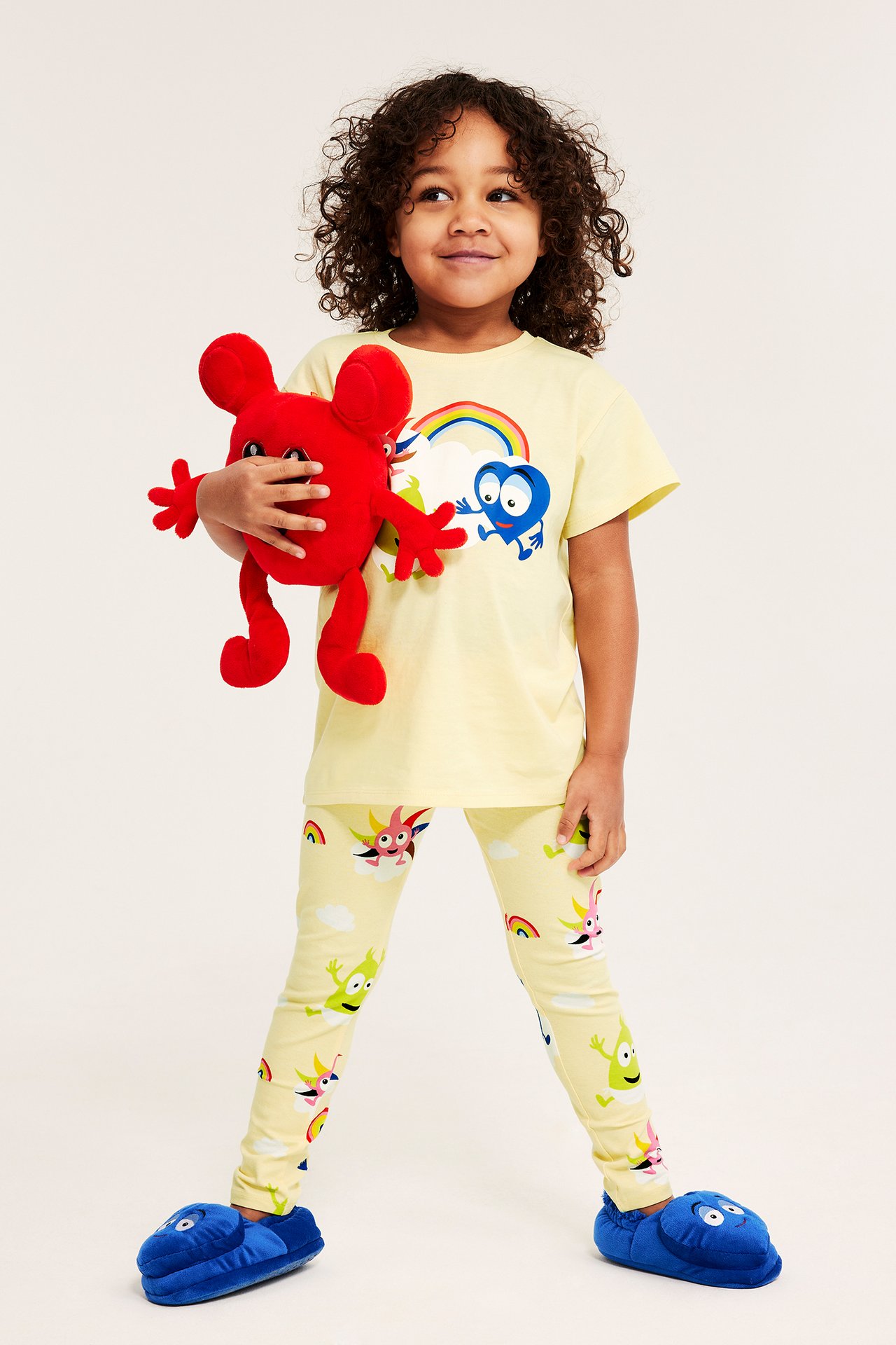 Edestä: Lapsi yllään vaaleanbeige Babblarna-kuvioitu t-paita, yhteensopivat legginsit, siniset tossut, pitäen punaista lelua.