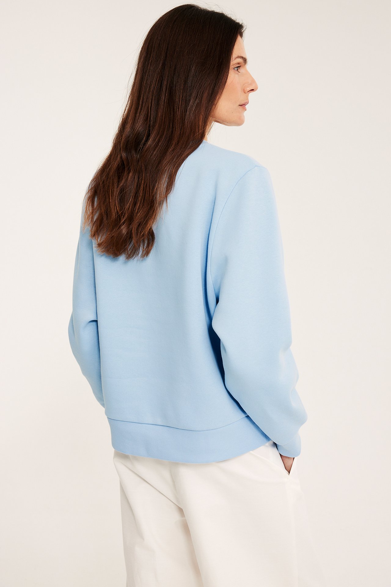 Widok z boku kobiety w jasnoniebieskiej, jednokolorowej bluzie z mieszanki bawełny i białych spodniach.