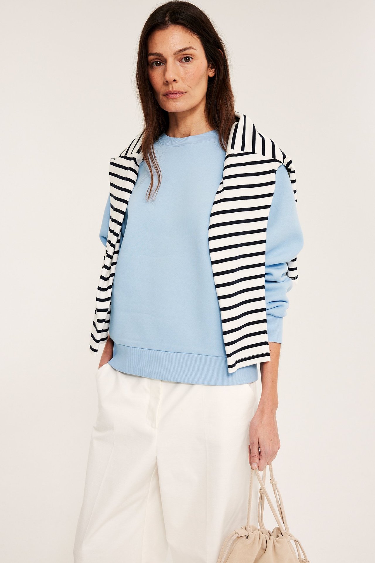 Frontutsikt: Kvinne i lys blå sweatshirt, hvite bukser, stripet topp over skuldrene, holder beige veske.