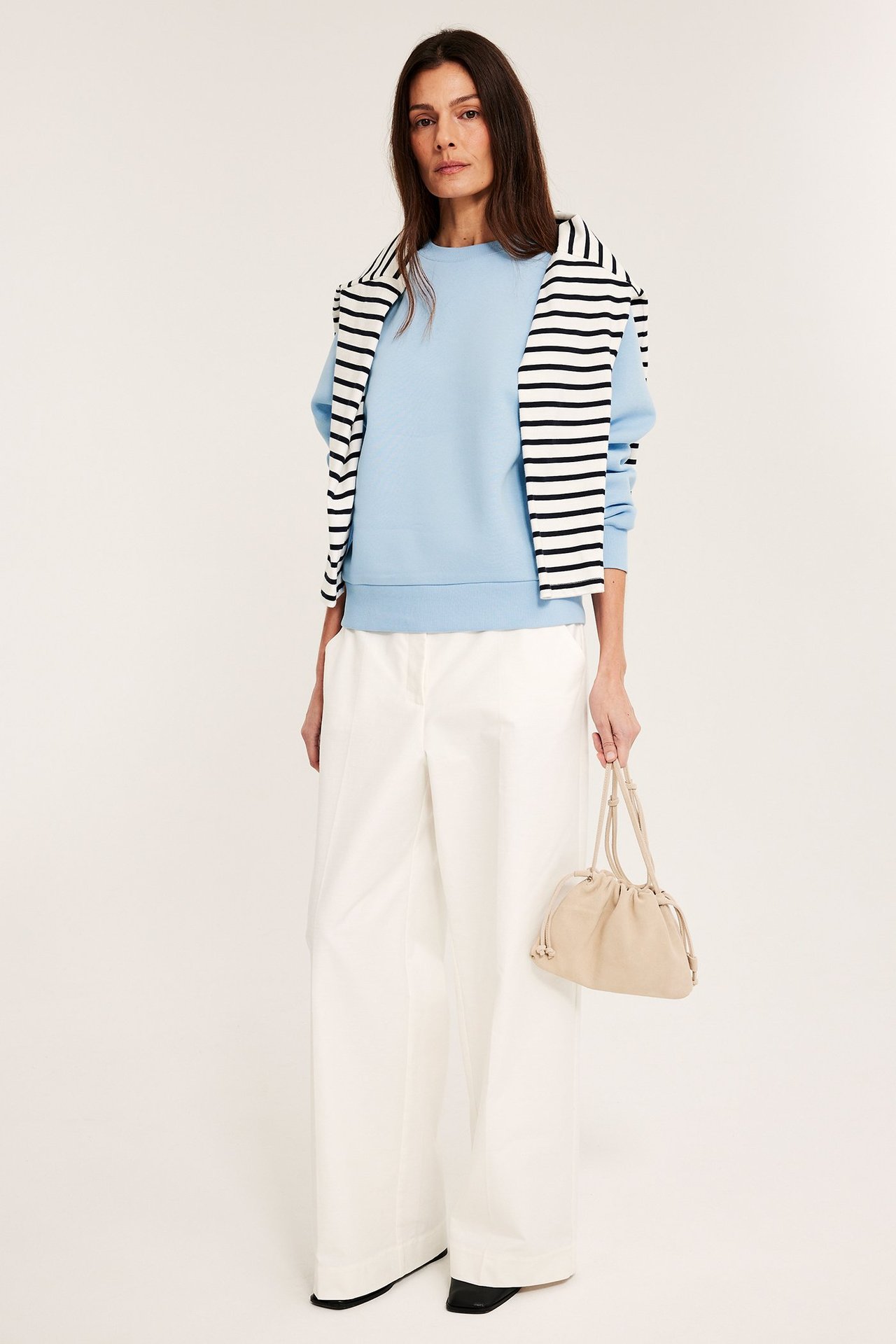 Widok z przodu: Kobieta w jasnoniebieskiej bluzie, białych spodniach, sweter w paski, z beżową torebką.