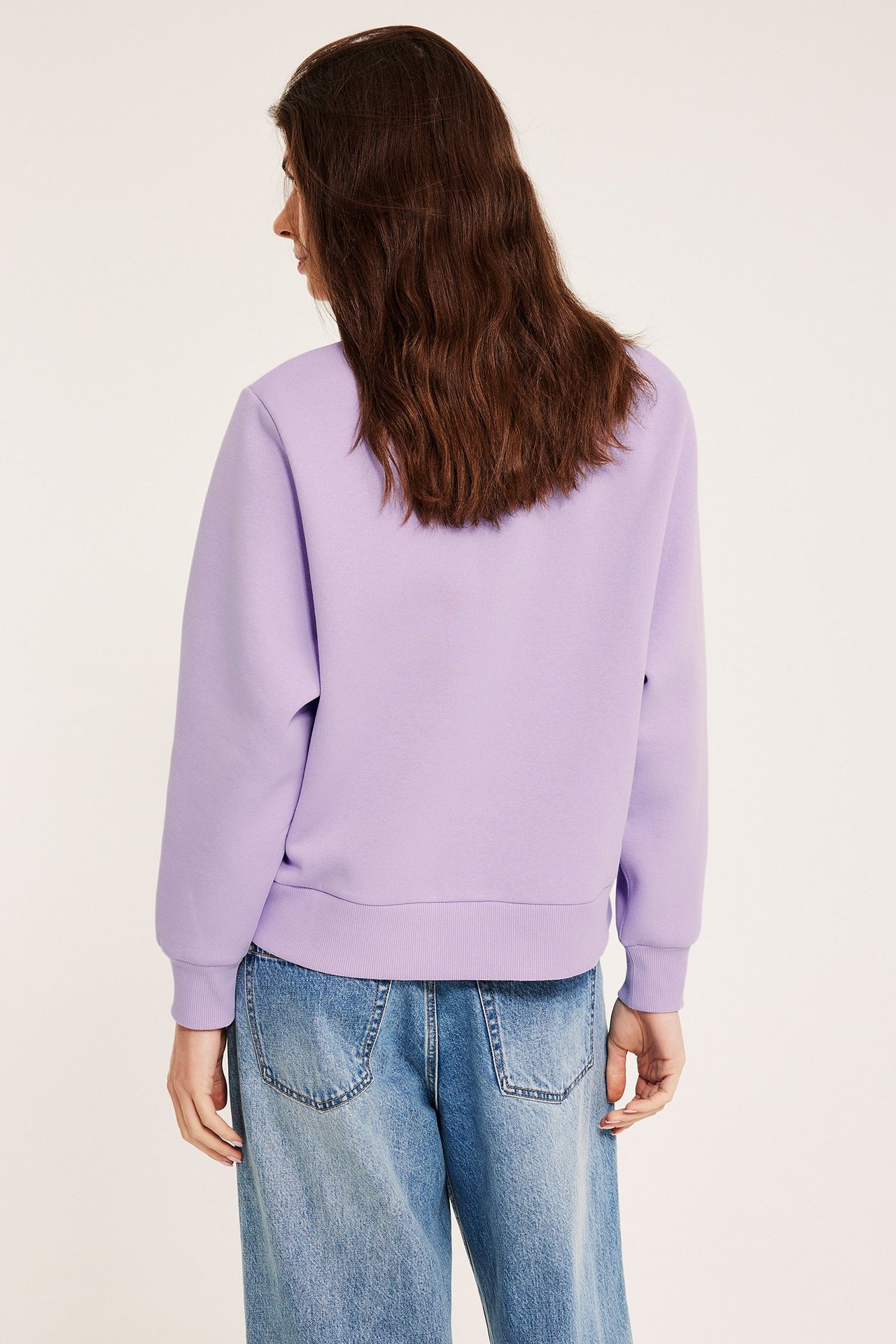 Naisen takakuva yllään vaaleanliila puuvillasekoitepusero resorisilla hihansuilla ja siniset, väljät farkut.