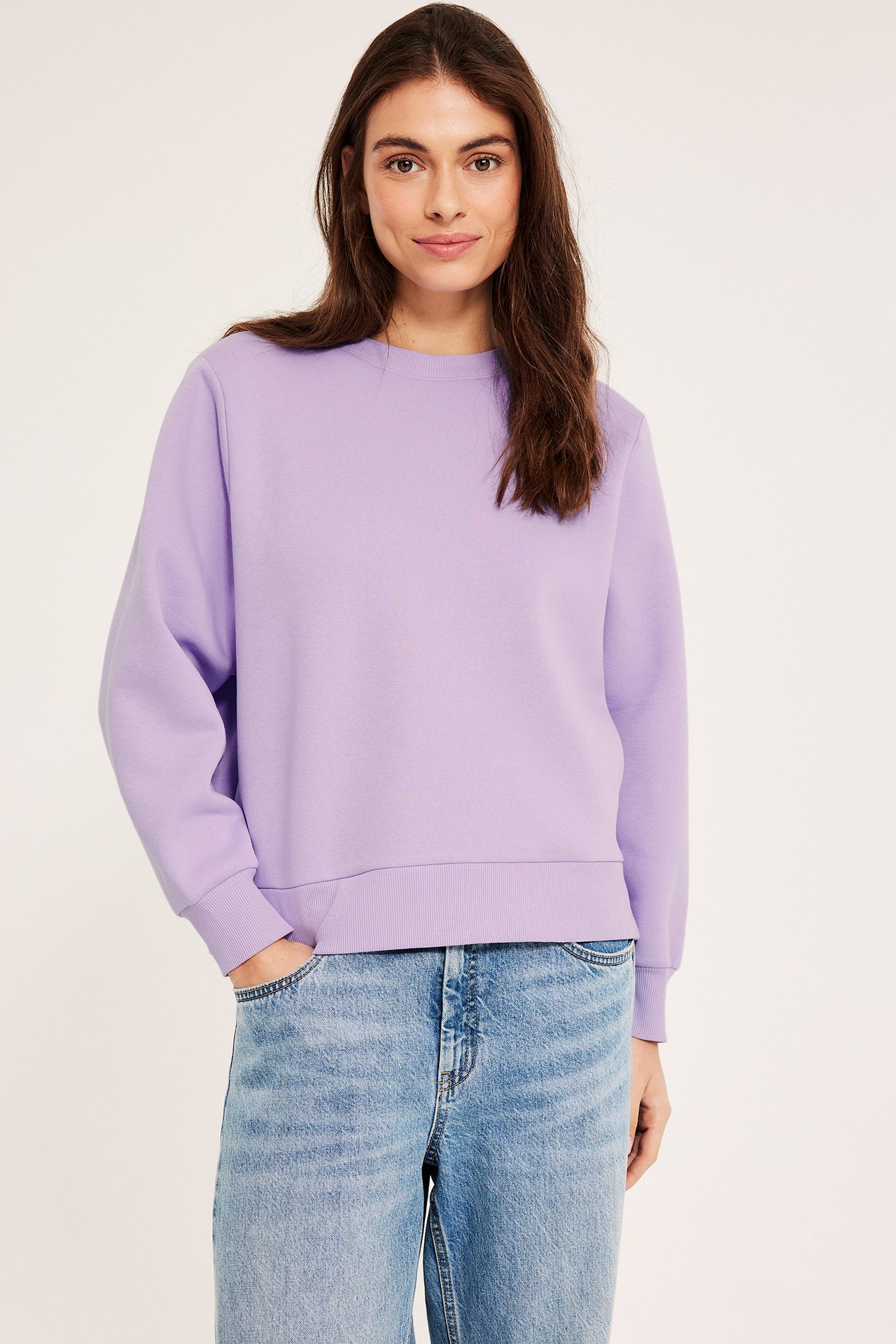 Edestä: nainen yllään vaaleanlila collegepaita resorireunoilla ja vaaleansiniset farkut.