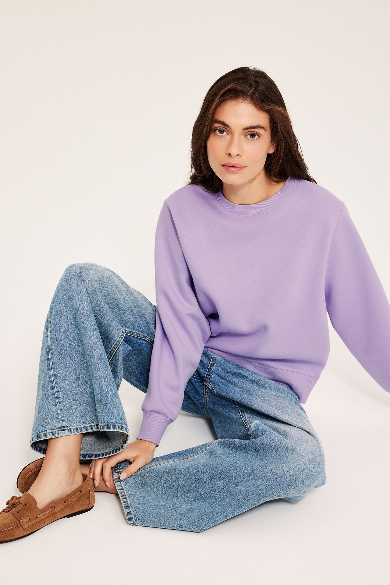 Edestäpäin, nainen yllään vaaleanliila collegepaita, leveälahkeiset farkut ja ruskeat mokkasiinit.