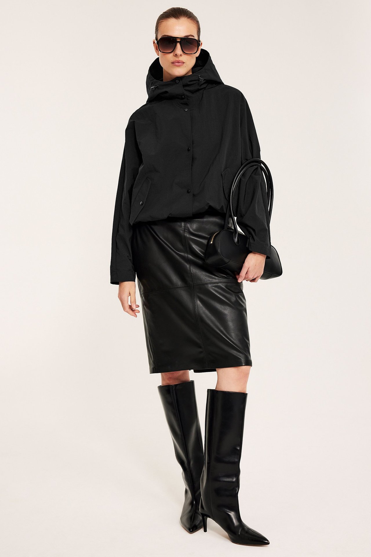 Widok z przodu: Kobieta w czarnej kurtce z kapturem, spódnicy z imitacji skóry, kozakach i torebce.