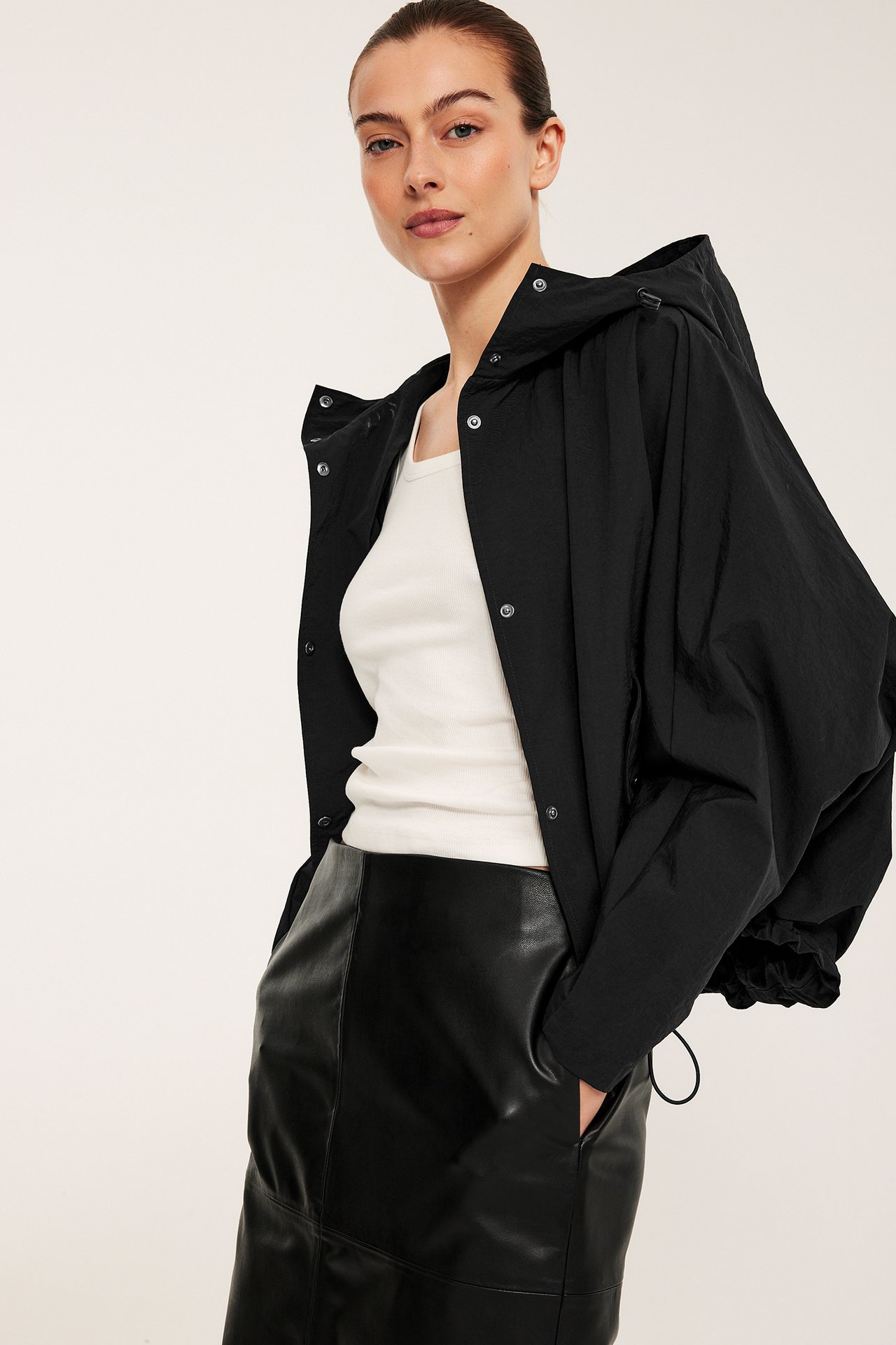 Widok z przodu: Kobieta w czarnej kurtce z kapturem na zatrzaski, białym topie i czarnej spódnicy.