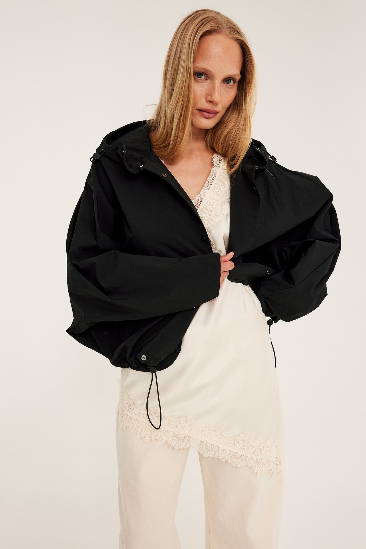 Front: Kvinne i svart, avslappet hettejakke over en hvit blonde-topp og lysebeige bukser.