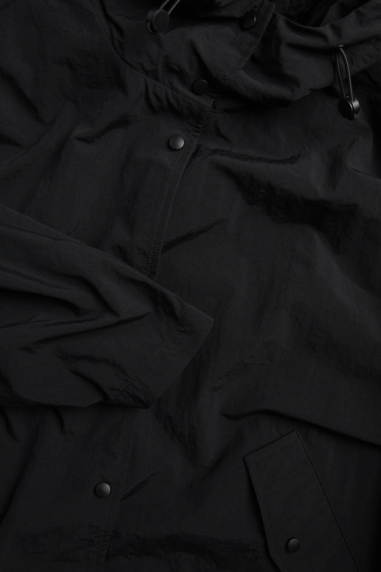 Närbild på en svart sportig jacka i polyamid med huva, dragsko och tryckknappsstängning.