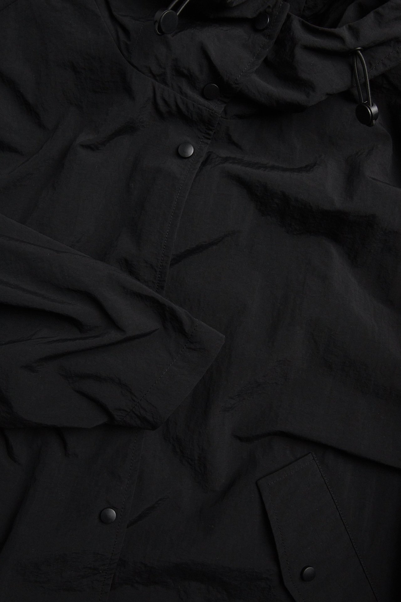 Närbild på en svart sportig jacka i polyamid med huva, dragsko och tryckknappsstängning.