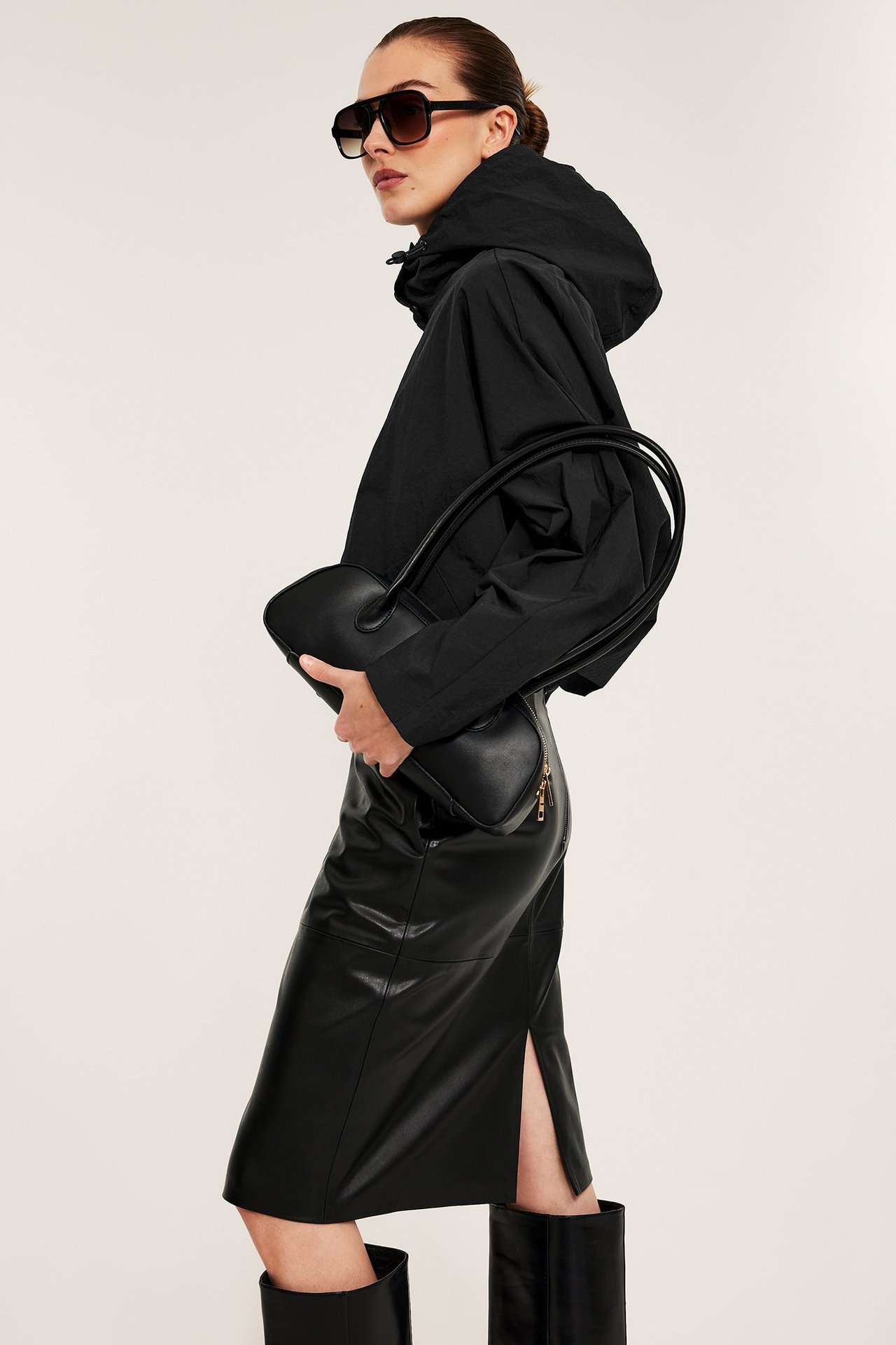 Widok z boku: kobieta w czarnej kurtce z kapturem, spódnicy midi ze sztucznej skóry, butach, okularach, z czarną torebką.