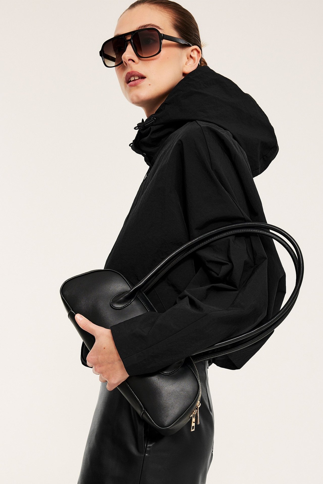 Sideprofil: Kvinne i svart avslappet hettejakke med snøring, holder svart håndveske og solbriller.