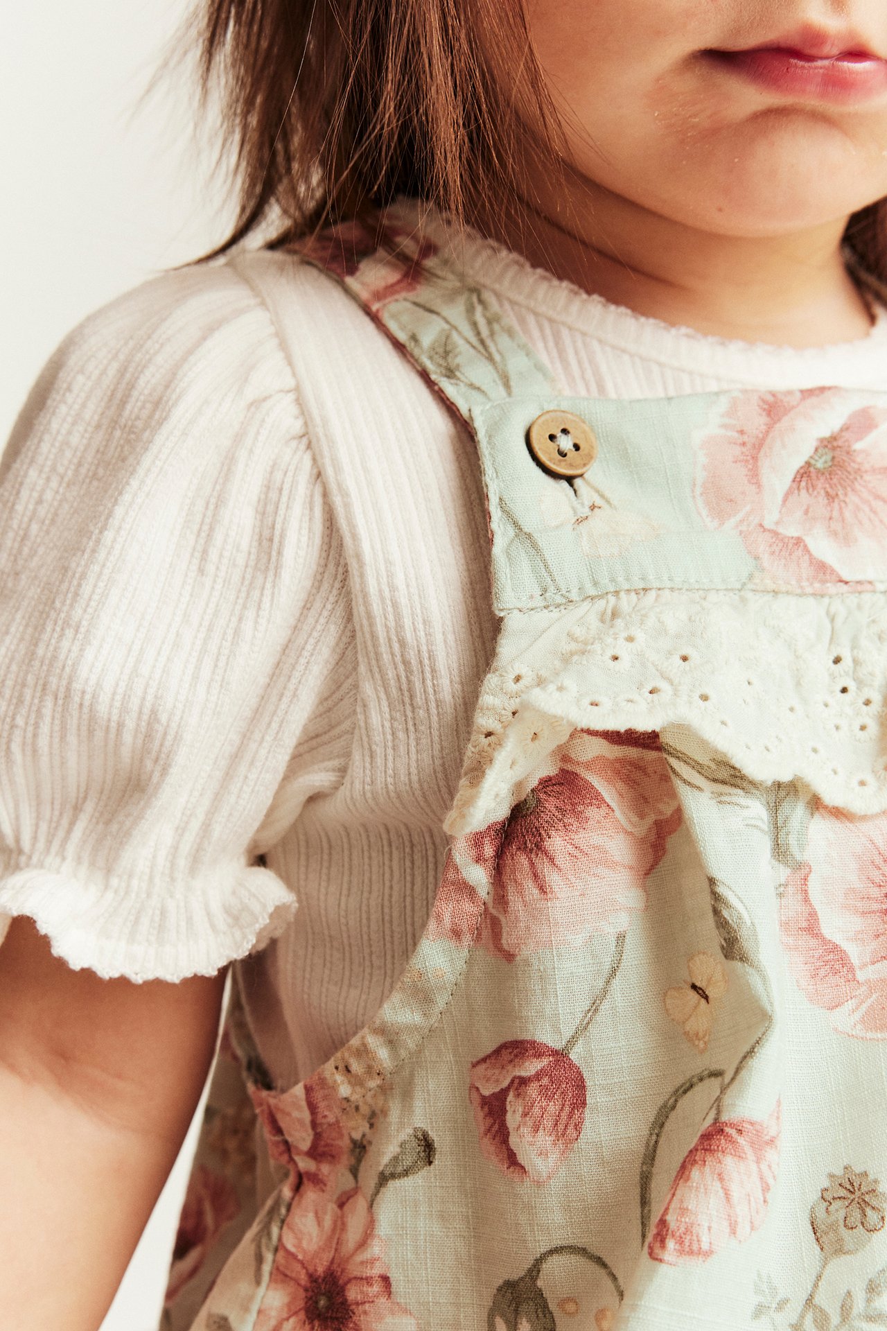 Zbliżenie dziecka w jasnozielonych ogrodniczkach w kwiaty z koronkową falbanką, na białym prążkowanym topie.