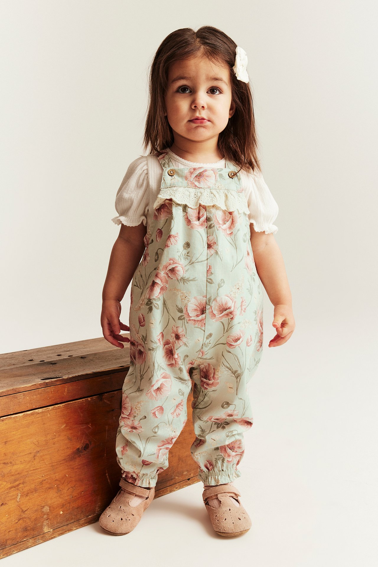 Etunäkymä lapsesta vaaleanvihreissä kukallisissa puuvillahaalareissa valkoisen ribbipaidan päällä, ruskeat kengät.