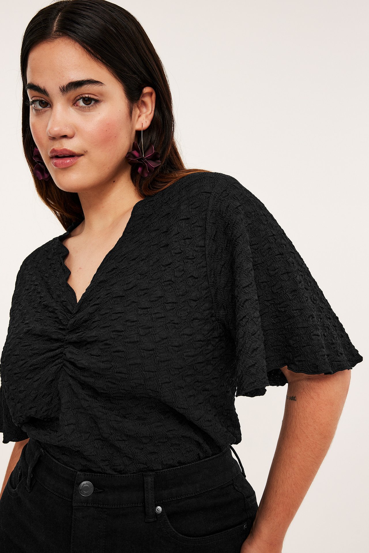 Framifrån: kvinna i svart strukturerad topp med rynkad front, volangärmar, svarta jeans och lila blomörhängen.