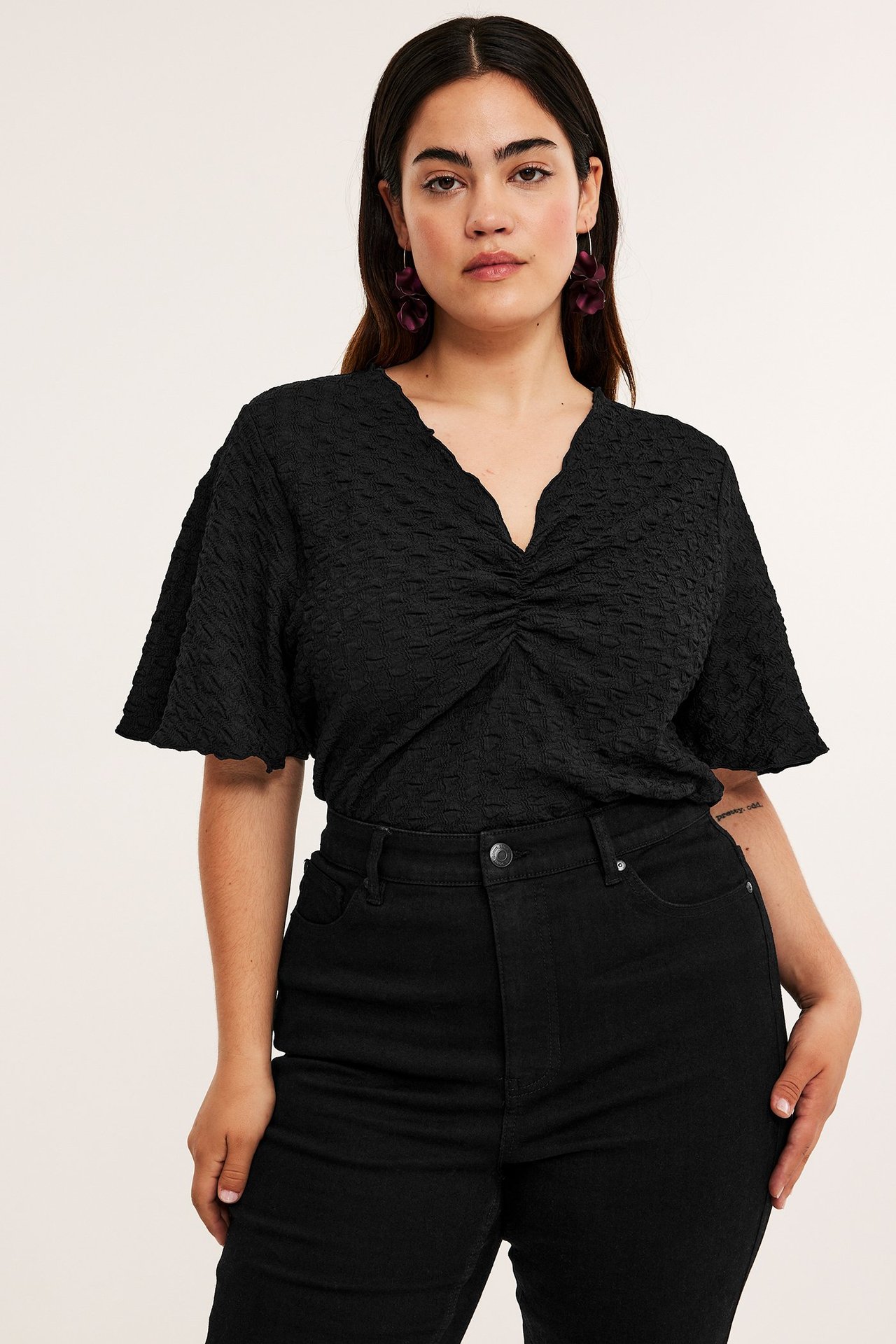 Widok z przodu: kobieta w czarnym teksturowanym topie z krótkim rękawem i marszczeniem, czarnych jeansach, fioletowych kolczykach.