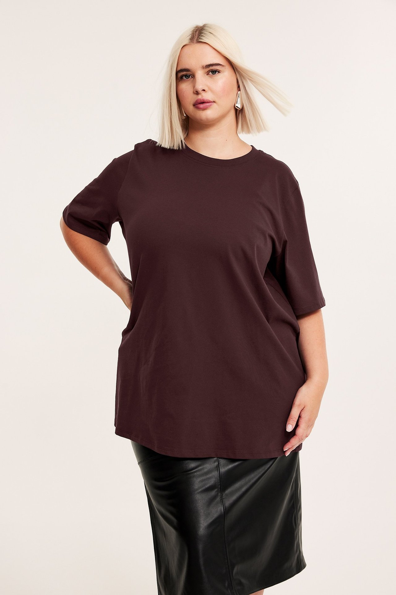 Widok z przodu kobiety w długim, oversize'owym bordowym bawełnianym T-shircie i czarnej spódnicy ze sztucznej skóry.