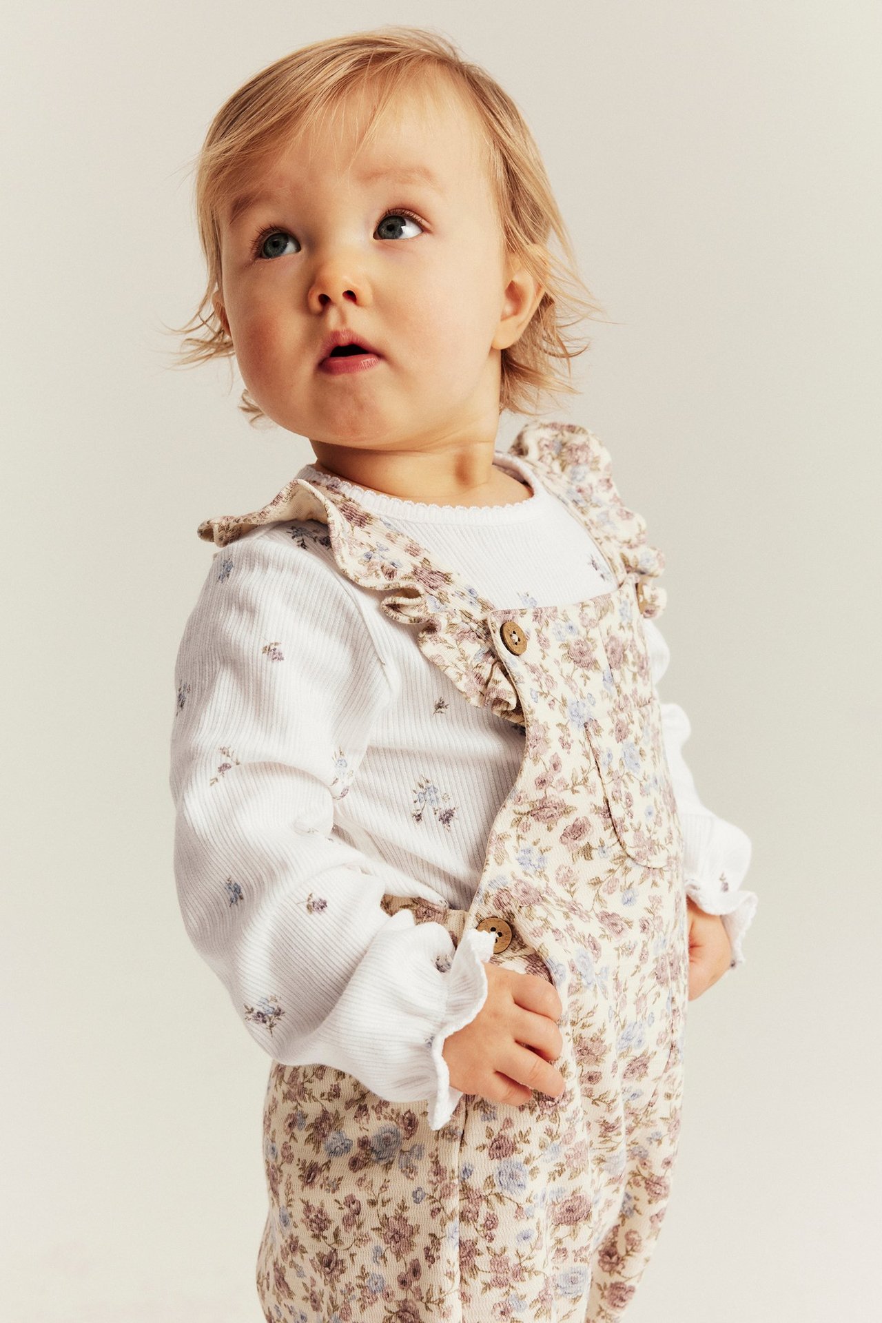 Frontvisning av baby i en offwhite blomstret ribbestrikket body med volangmansjetter, under en blomstret overall.