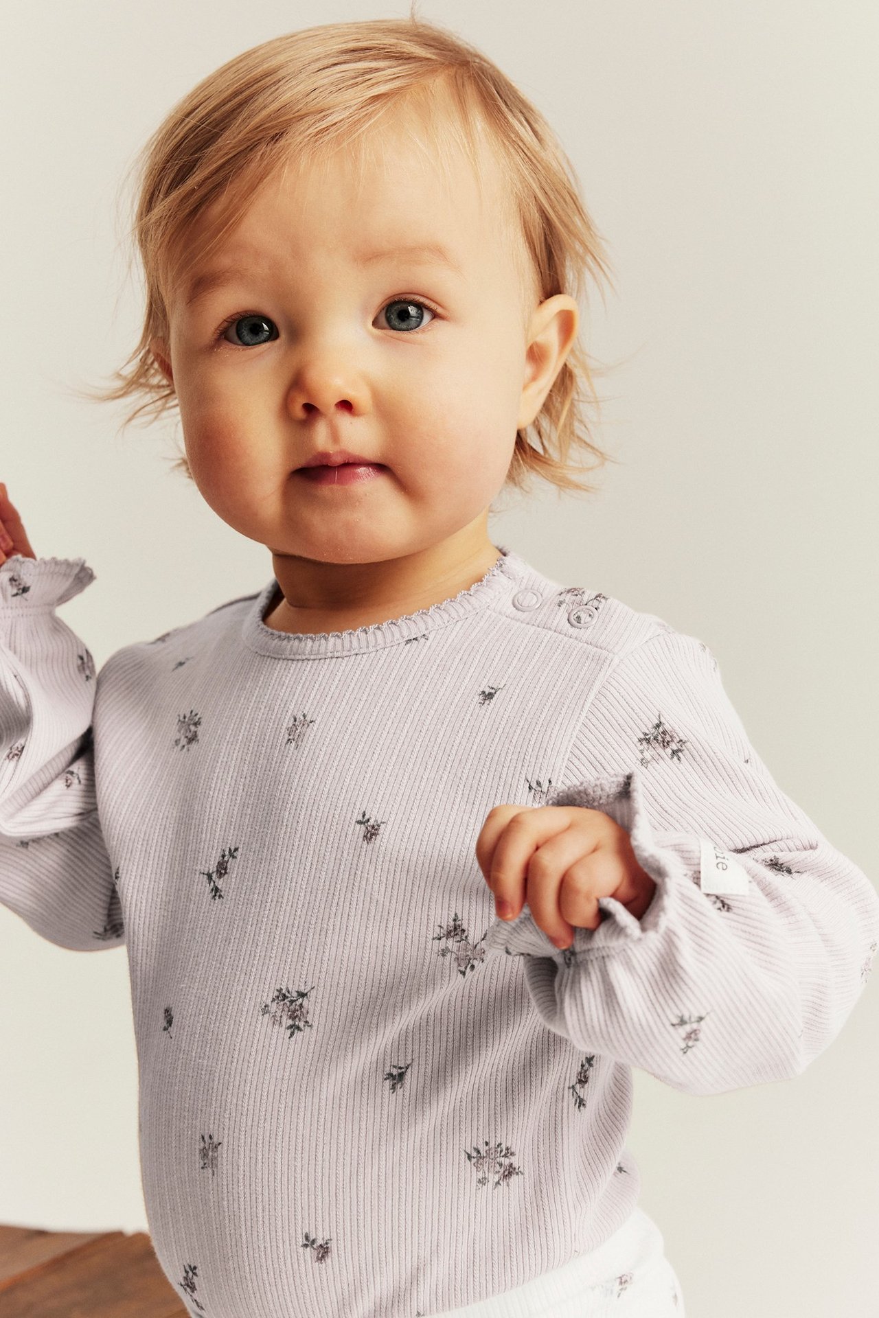 Nærbilde forfra av en baby i lys lilla ribbet body med blomstermønster, volanger og trykknapper.