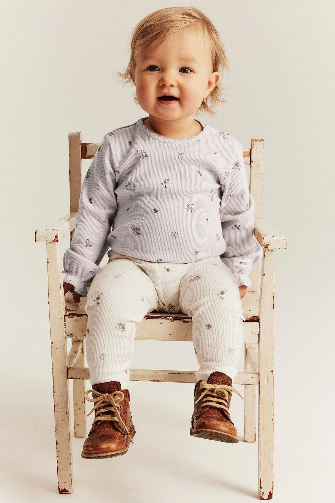 Front: baby i lys lilla ribbet body med blomster, kremfargede blomstrete leggings og brune snørestøvler.