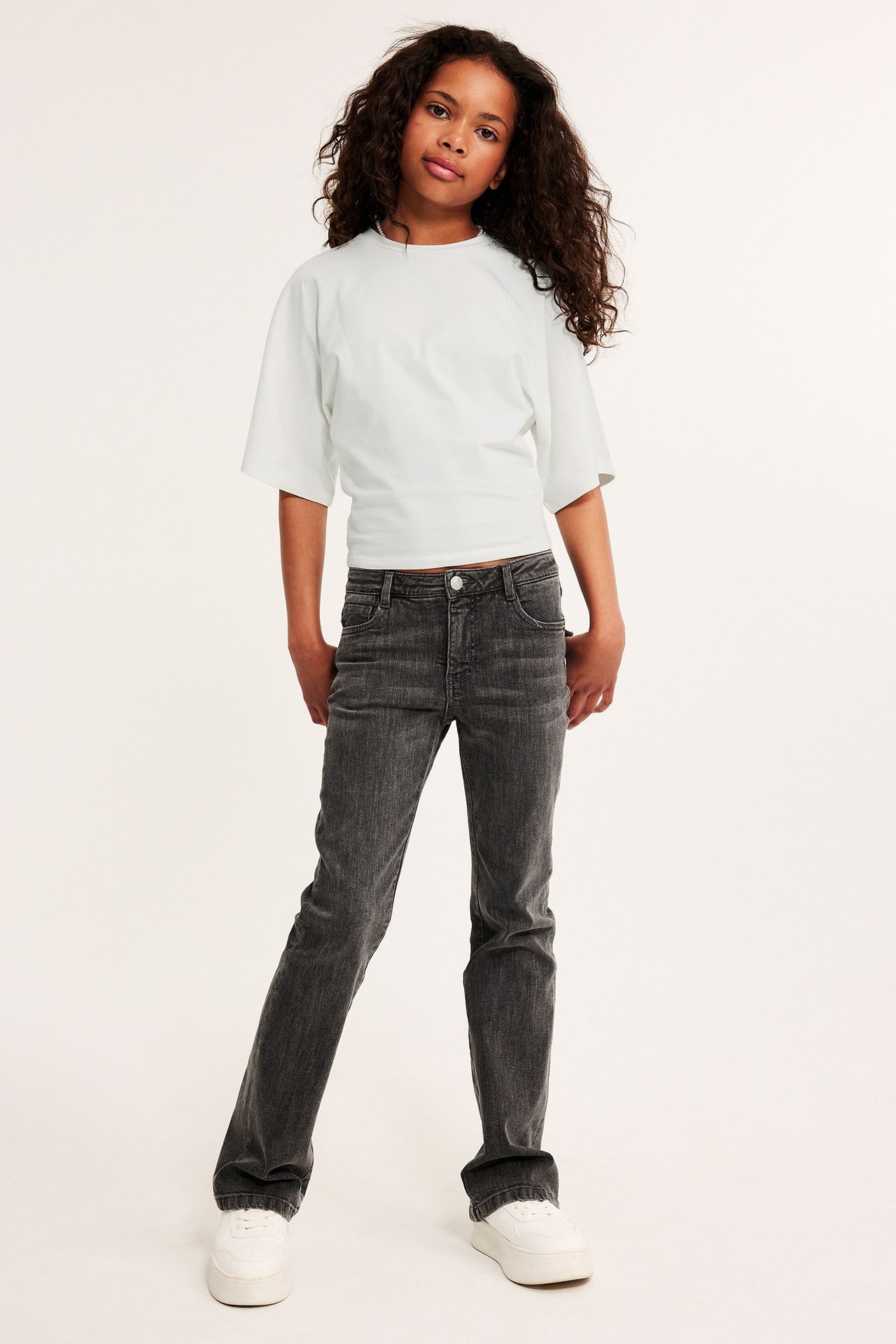 Framifrån: en tjej bär en offwhite topp med fjärilsärmar, mörkgrå jeans och vita platåsneakers.