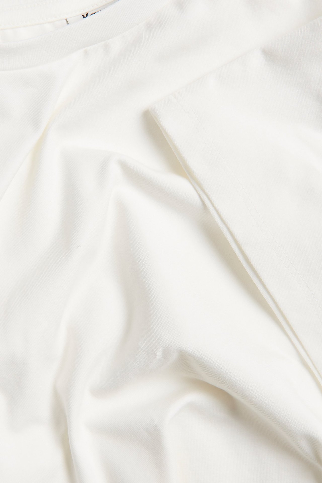 Zbliżenie na gładki, złamanej bieli materiał topu z mieszanki bawełny z delikatnymi fałdami.