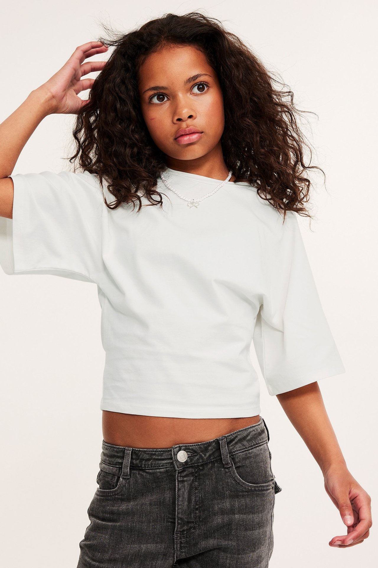 Frontvy av ett barn i en offwhite kort topp med fjärilsärmar, mörkgrå jeans och ett pärlhalsband.