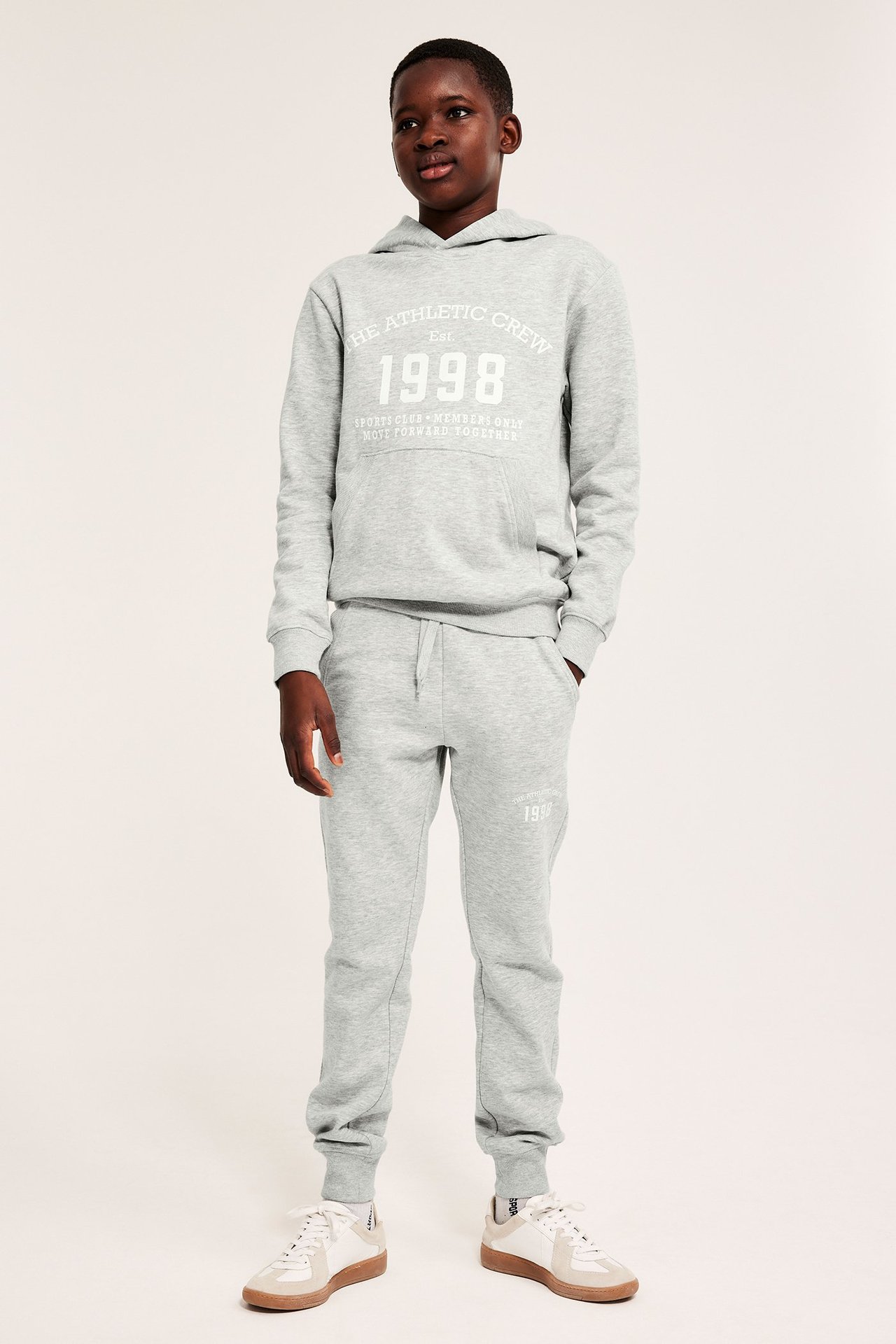 Framifrån: Barn bär gråmelerade mjukisbyxor med tryck, matchande huvtröja och vita sneakers.