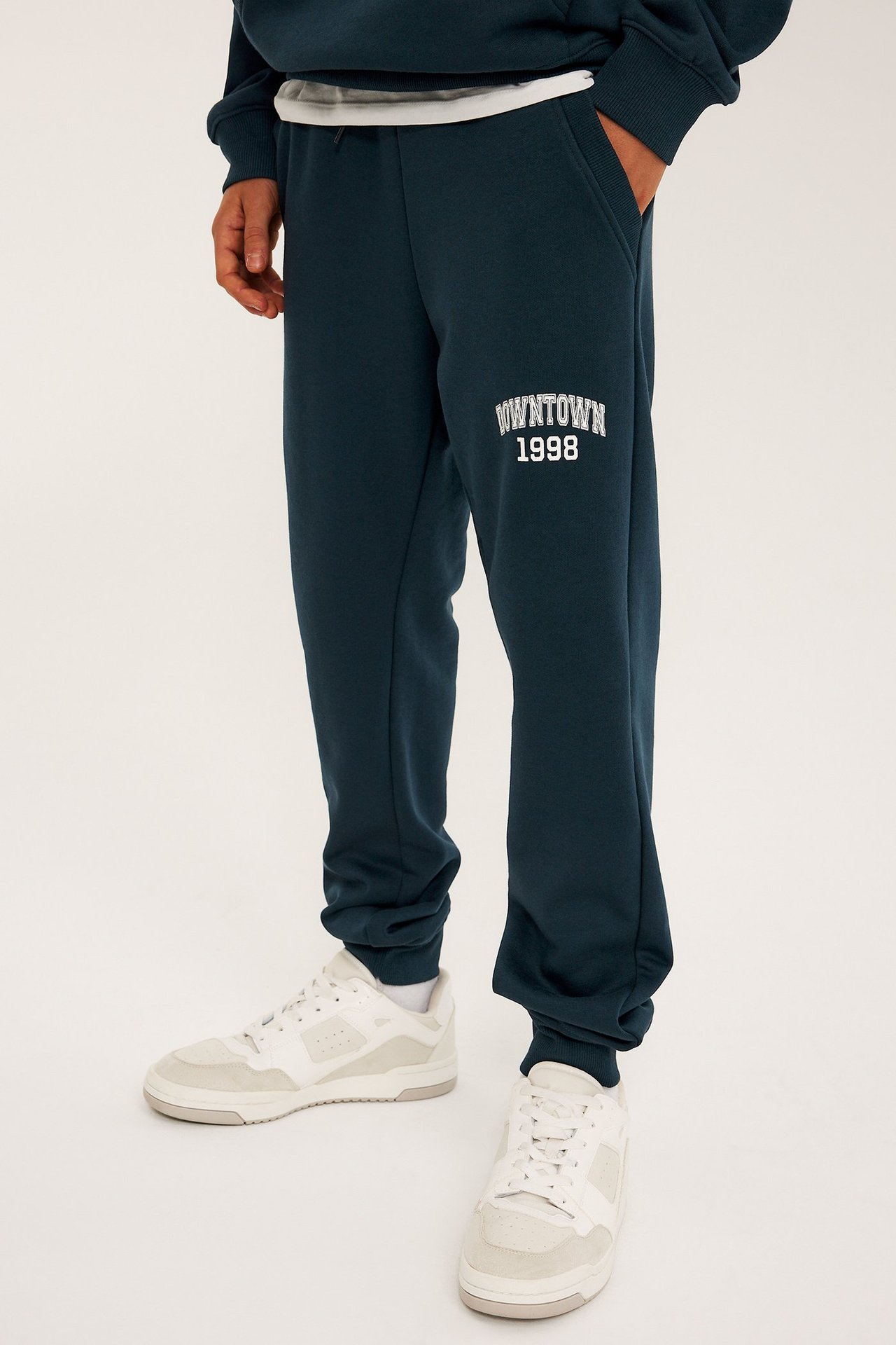 Edestä: Lapsi sinisissä puuvillasekoitecollegehousuissa 'DOWNTOWN 1998' -printillä, valkoiset tennarit, sininen huppari.