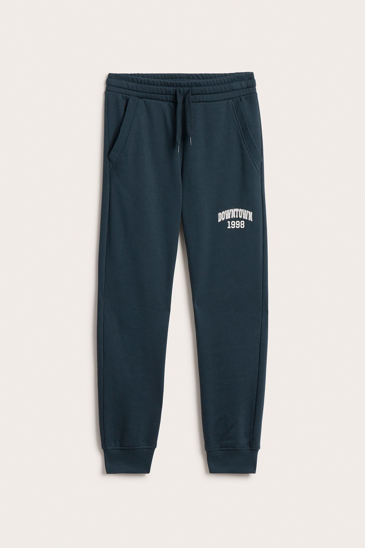 Edestä: Tummansiniset lasten collegehousut joustovyötäröllä, lahkeensuissa, valkoinen 'DOWNTOWN 1998' -printti.