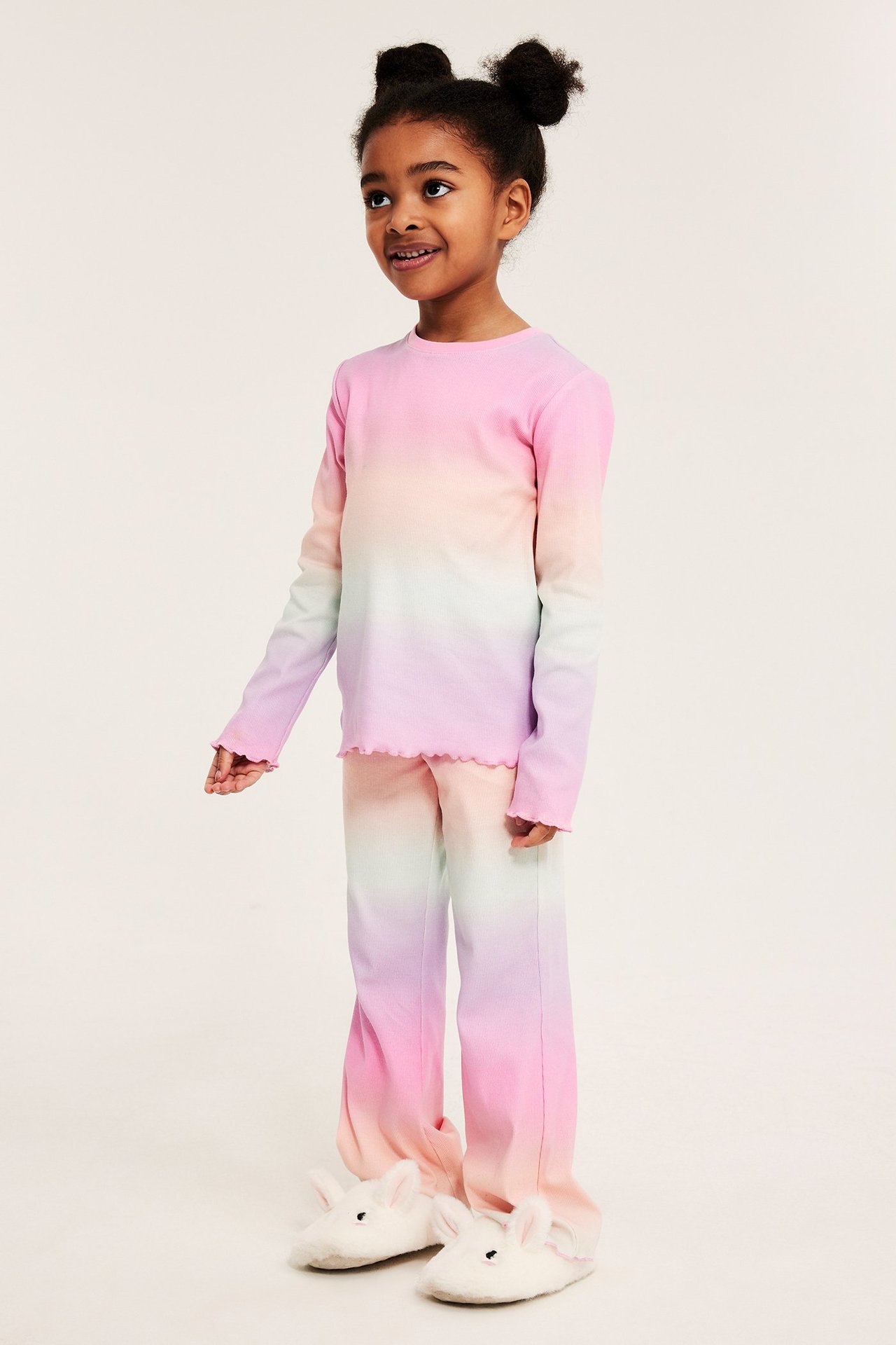 Widok z przodu: Dziecko w prążkowanej piżamie w odcieniach różu, zieleni, fioletu, w białych kapciach króliczkach.