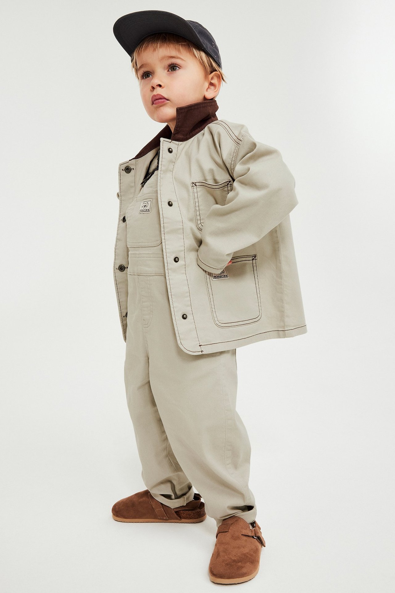 Edestä: Lapsi yllään beige twill-takki, beiget haalarit, tumma lippalakki ja ruskeat pistokkaat.