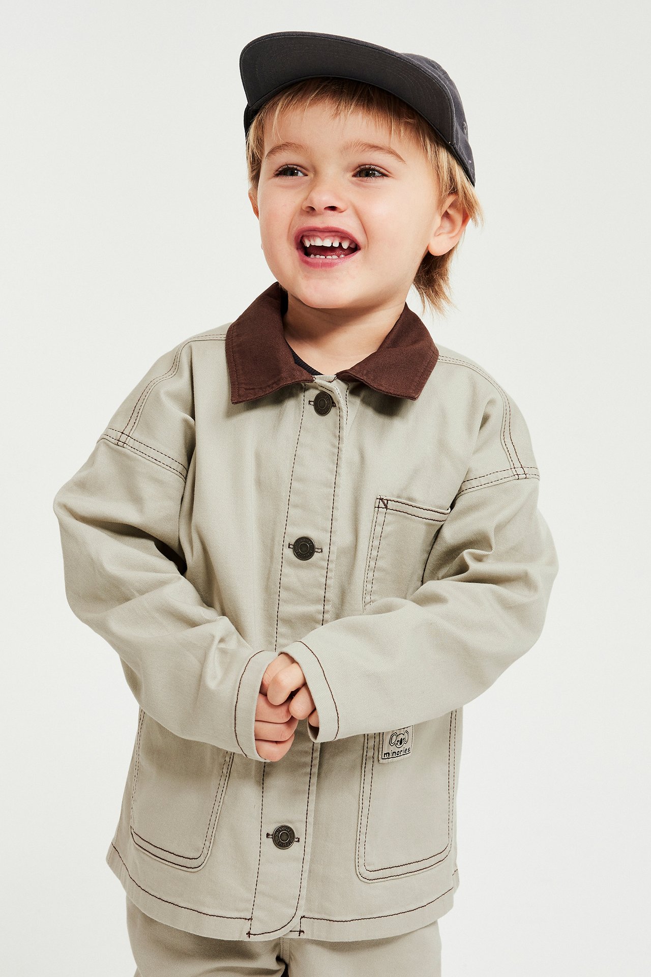Framifrån: Ett barn bär en beige twilljacka med brun krage, mörka knappar och en mörkgrå keps.