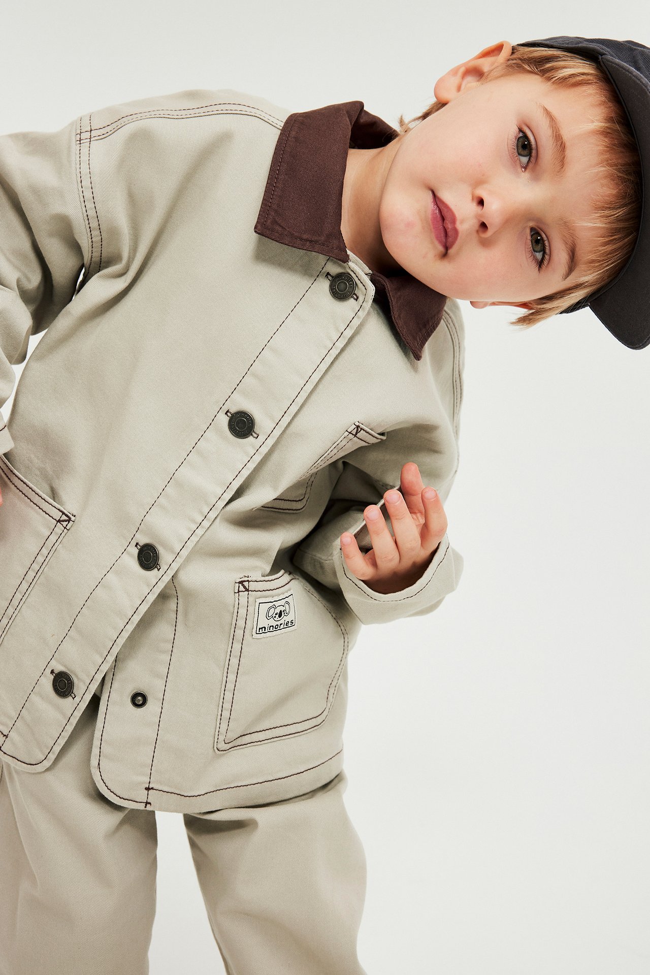 Frontvy av ett barn i beige twilljacka med brun krage, mörka knappar och mörk keps.