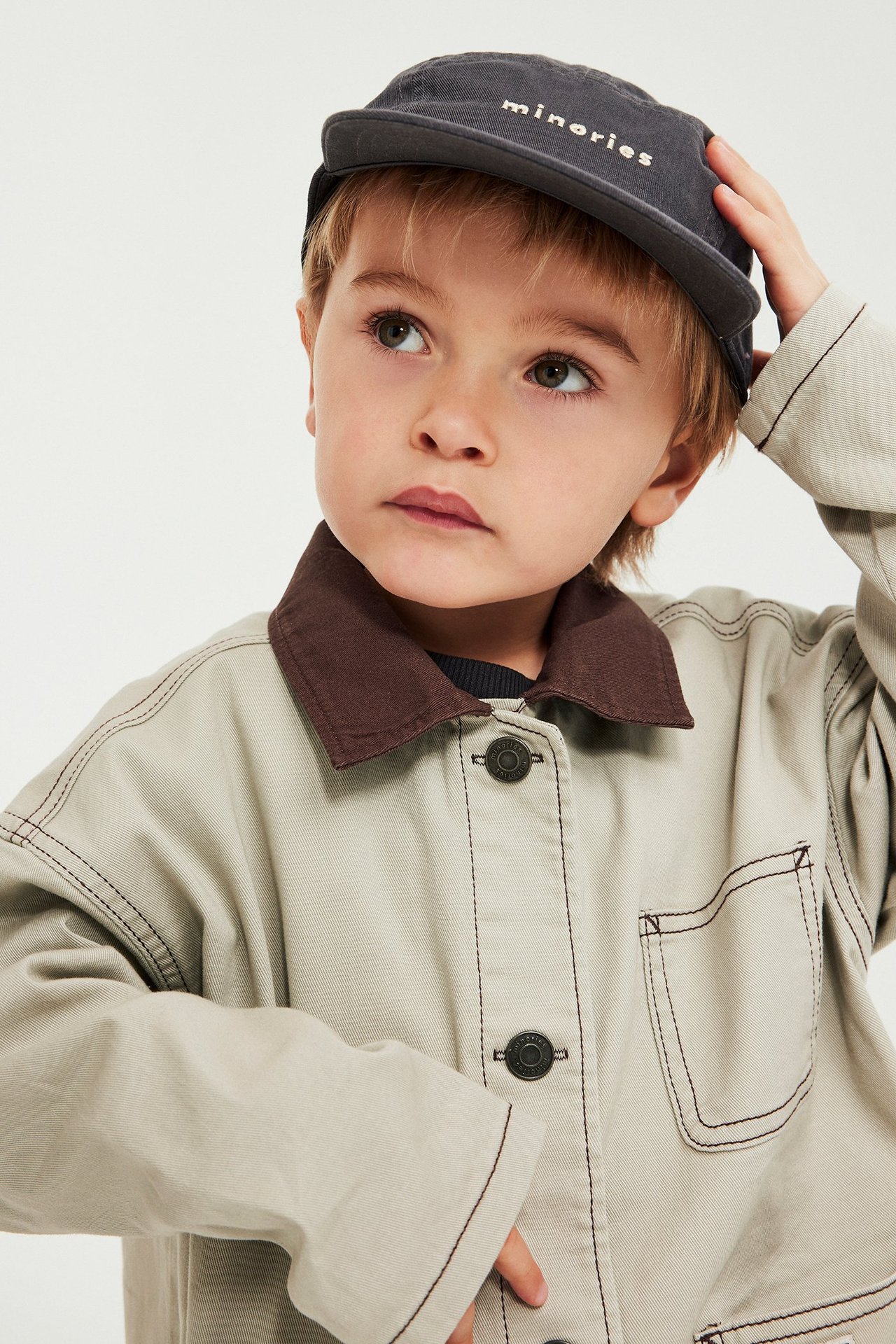 Närbild framifrån på ett barn i beigefärgad twilljacka med brun krage och mörkgrå keps.