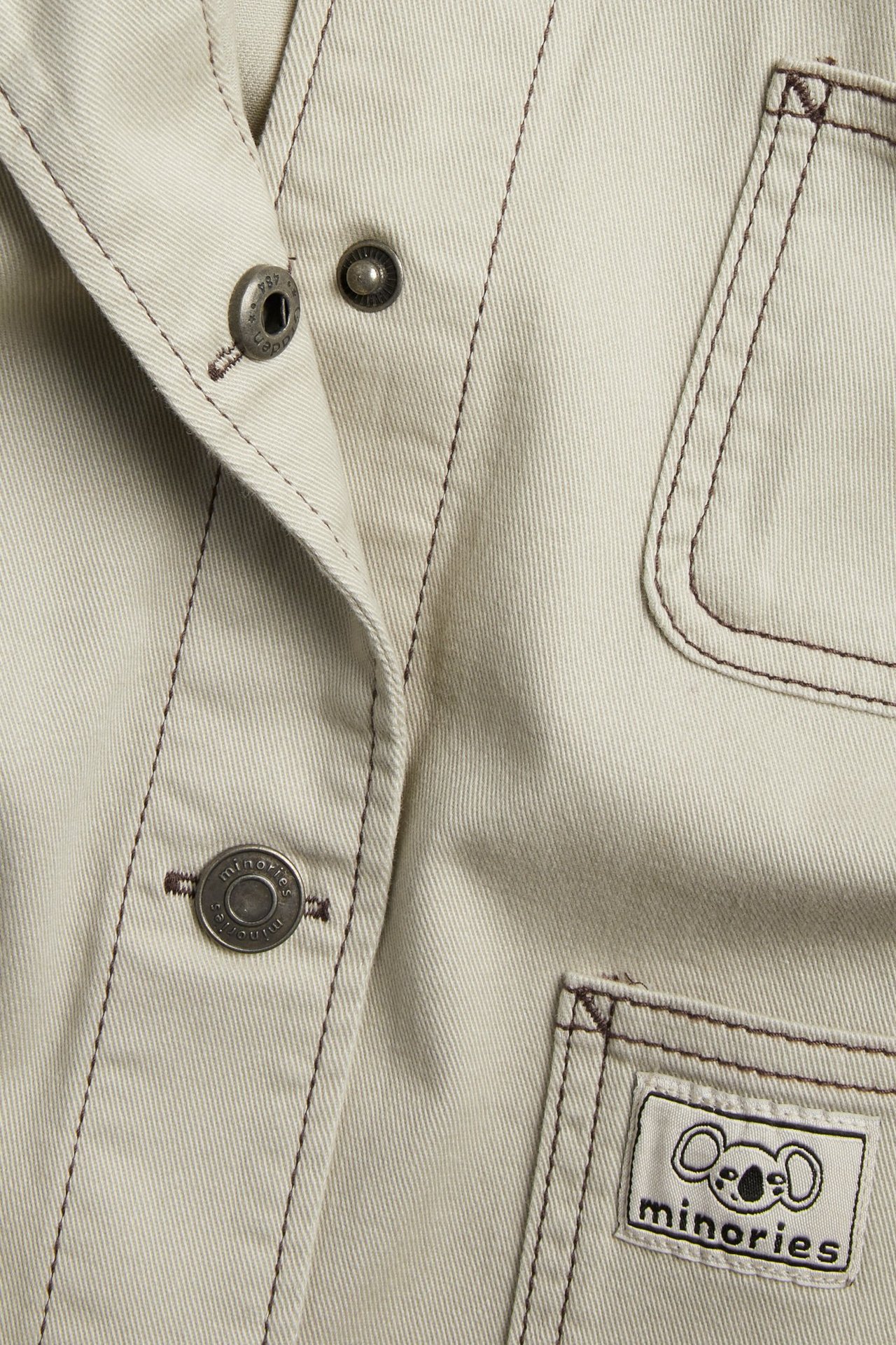 Zbliżenie beżowej kurtki twillowej z ciemnobrązowymi szwami, metalowymi napami i metką 'minories'.