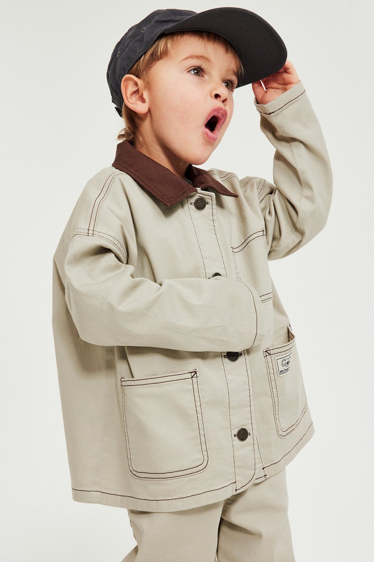 Edestä: Lapsi beigessä puuvillatvillitakissa ruskealla kauluksella, tummilla napeilla, beigellä housulla ja tummalla lippalakilla.