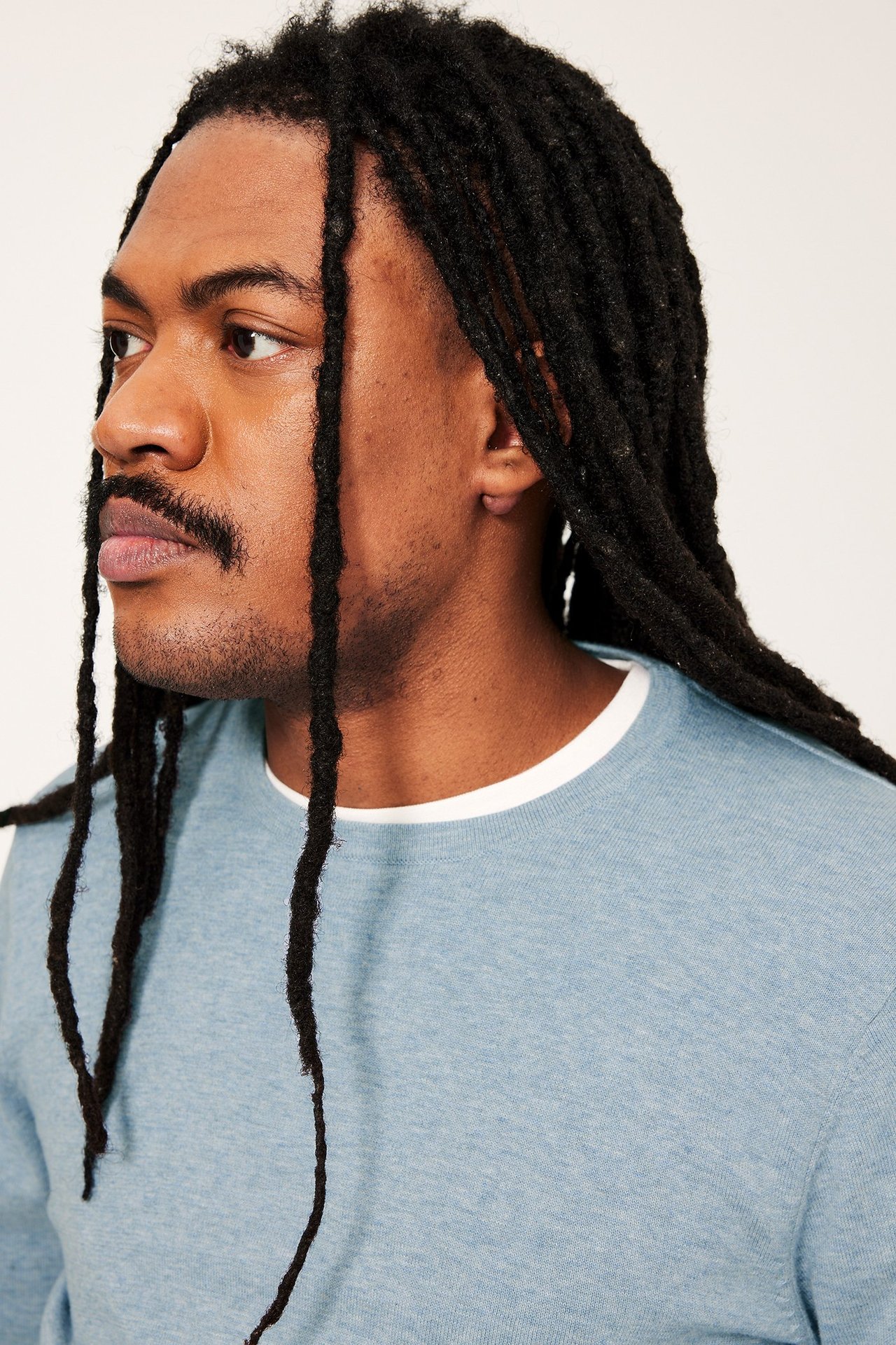 Nærbilde av mann i sidestilling, iført lyseblå melert finstrikket bomullsgenser og hvit t-skjorte.