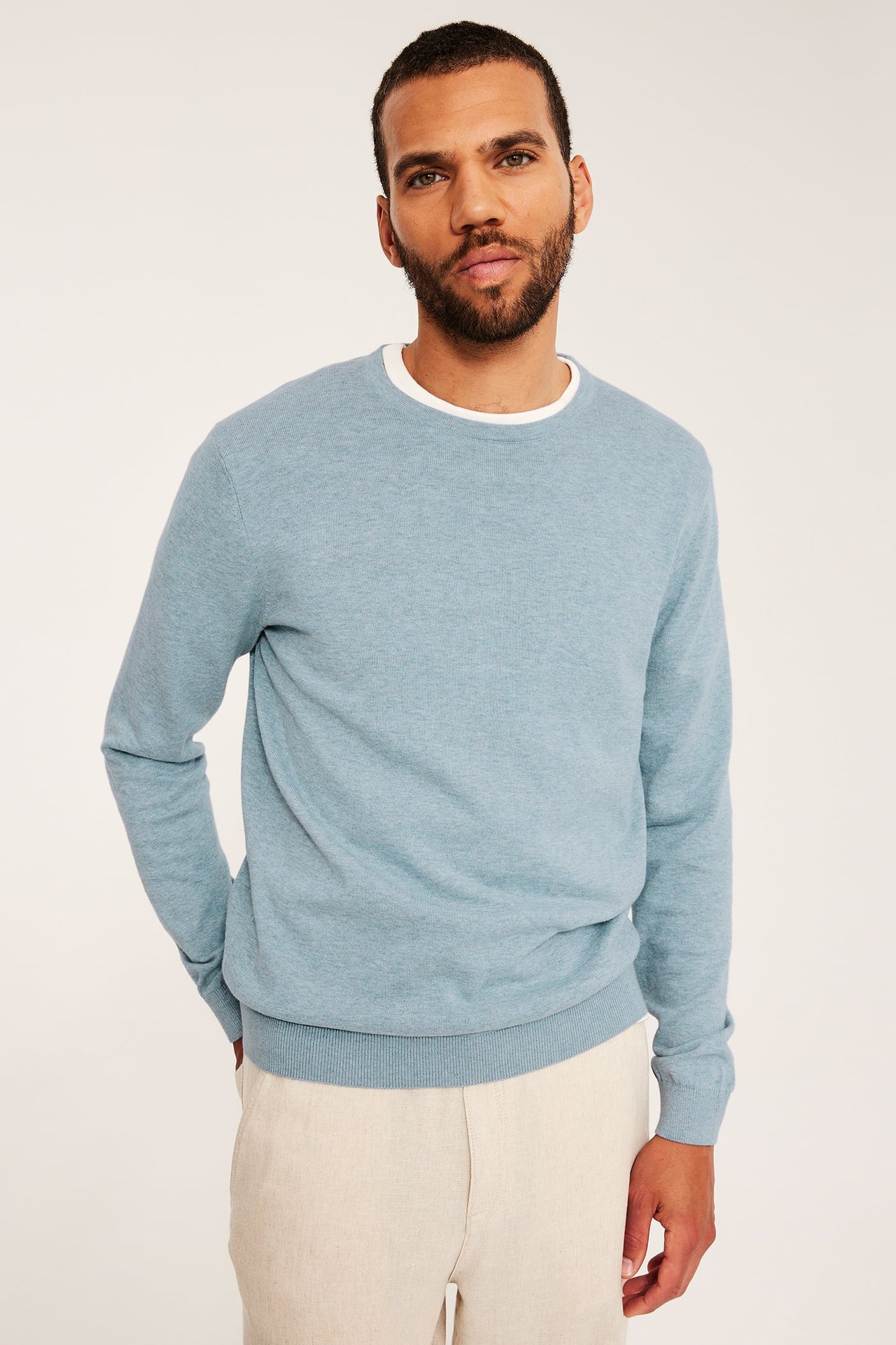 Frontvy av en man i ljusblå melerad finstickad bomullströja, vit t-shirt och ljusbeige byxor.