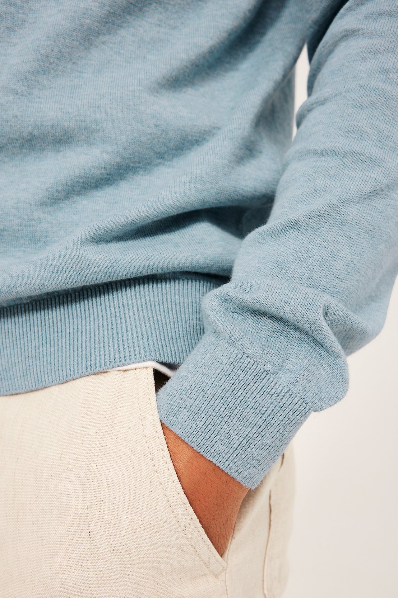 Närbild från sidan: Man bär en ljusblå melerad finstickad bomullströja och ljusbeige byxor.