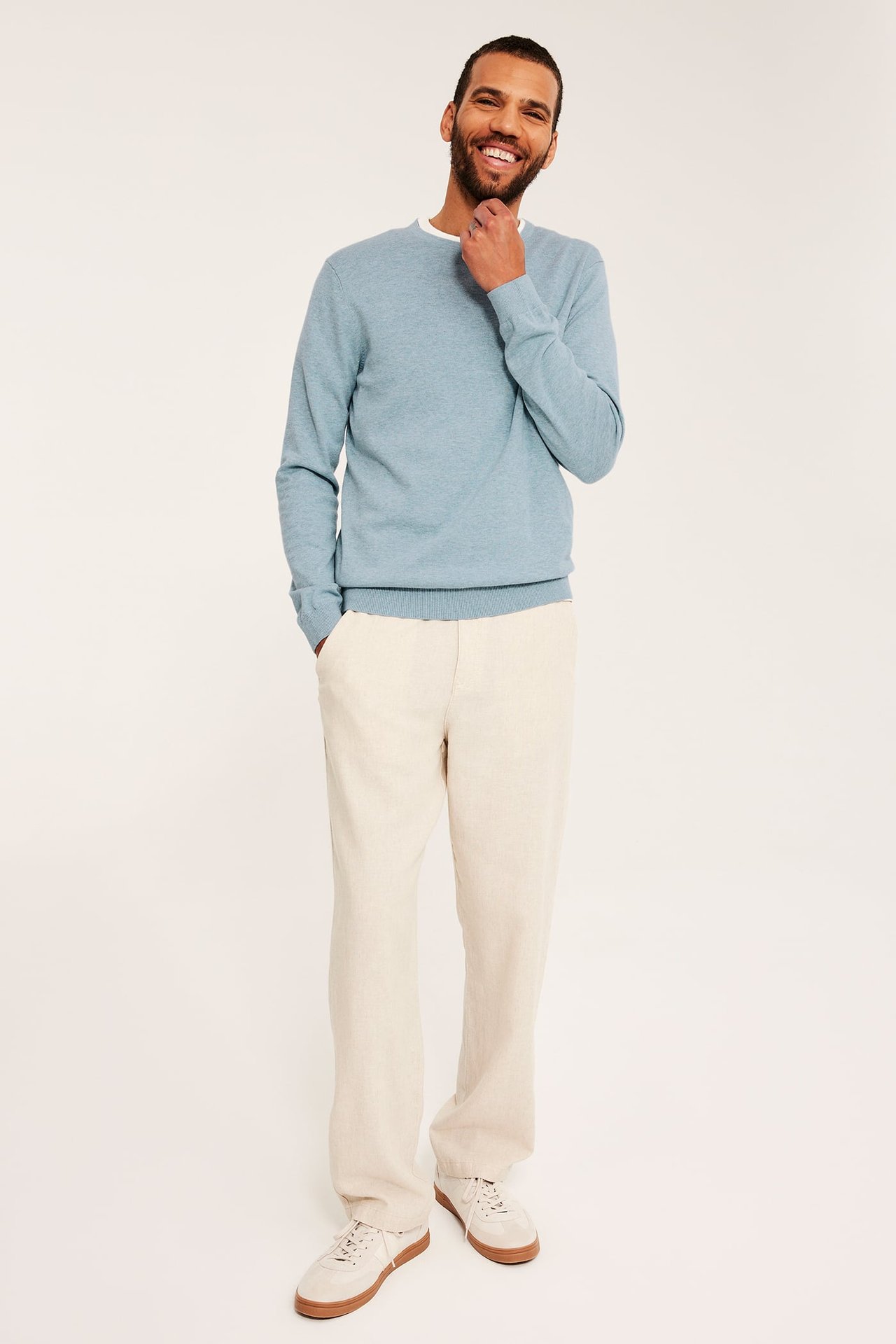 Frontvisning: Mann har på seg lys blå melert finstrikket genser over hvit t-skjorte, beige bukse og joggesko.
