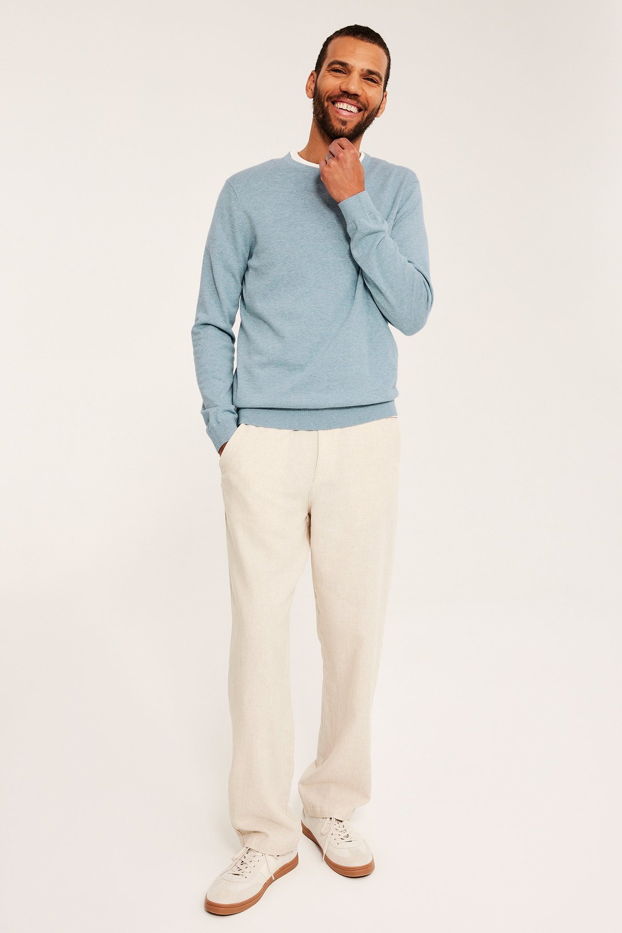 Frontvisning: Mann har på seg lys blå melert finstrikket genser over hvit t-skjorte, beige bukse og joggesko.