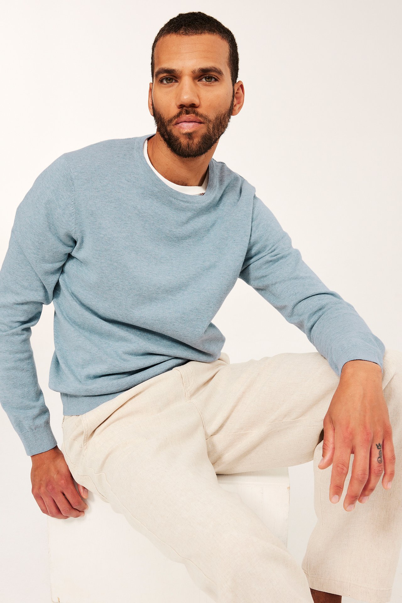 Edestä näkymä miehestä yllään vaaleansininen meleerattu neulepusero ja beiget housut.