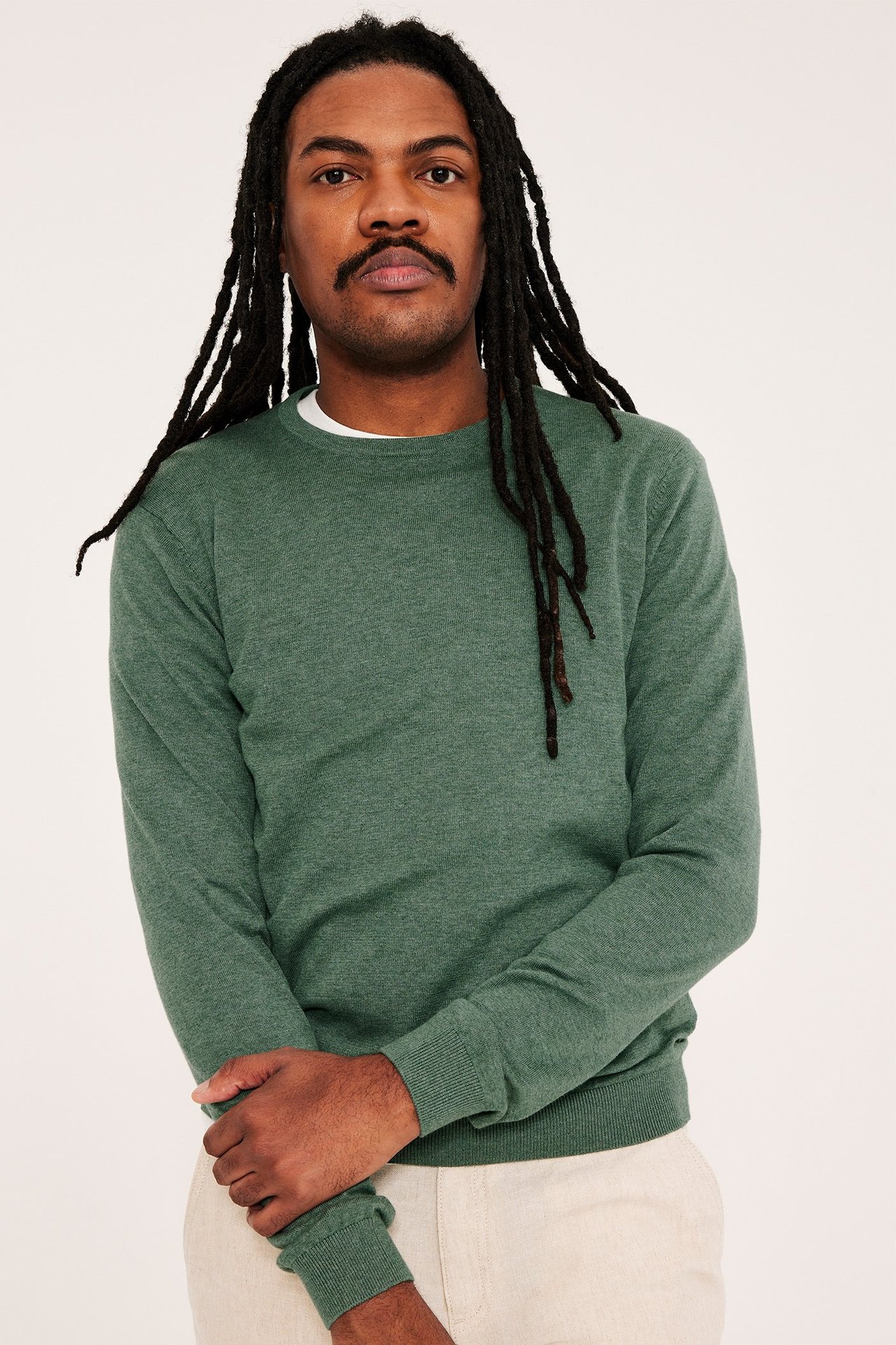 Framifrån, man i melerad grön finstickad bomullströja över vit t-shirt och ljusbeiga byxor.