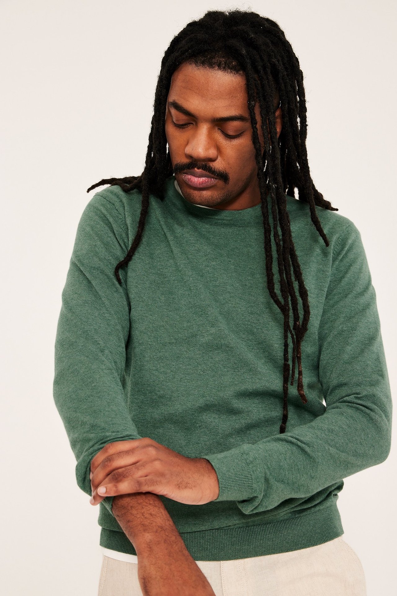 Widok z przodu mężczyzny w zielonym melanżowym cienkim swetrze bawełnianym, białym t-shircie i jasnobeżowych spodniach.